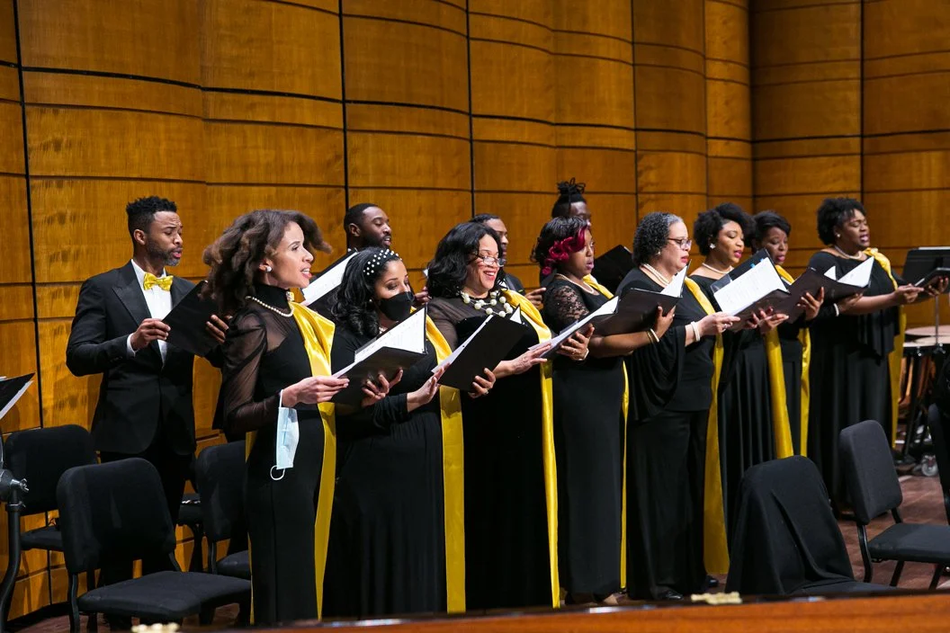 Choir of men and women singing in a concert hall, wearing black outfits with yellow sashes, holding sheet music.