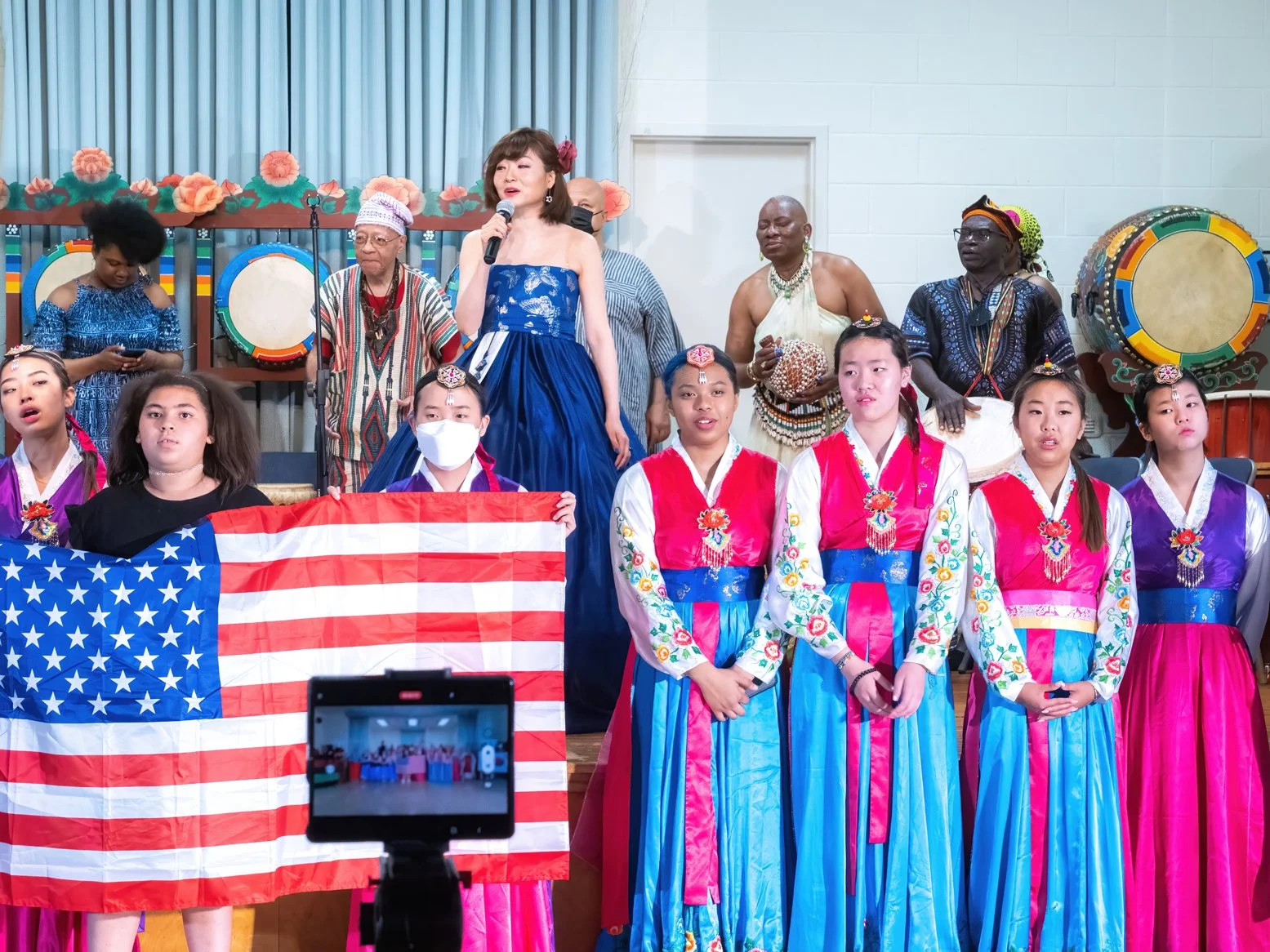 A diverse group of children and women on stage, some wearing traditional Korean hanbok, performing or participating in a cultural event. A woman in a blue dress is speaking into a microphone, while others hold a U.S. flag and traditional drums in the