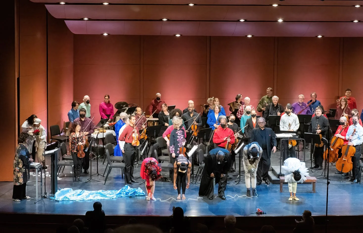 An orchestra on stage bowing after a performance, with musicians and crew members present, some wearing masks.