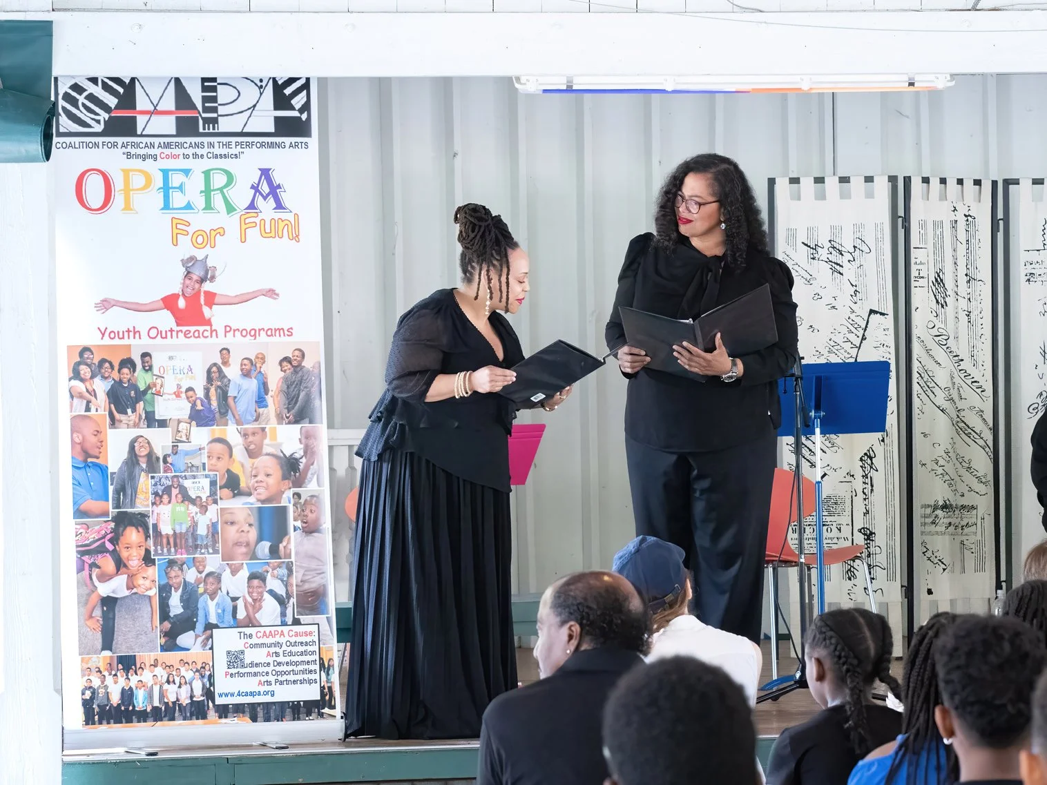 Two women in black attire standing on a stage, holding documents, in front of an audience at an event promoting youth outreach programs for opera music.
