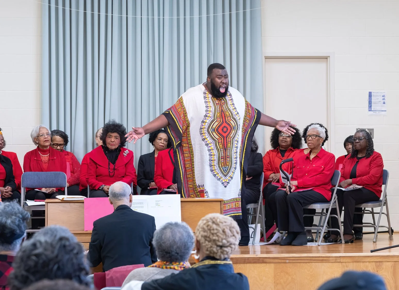 A man dressed in a colorful dashiki speaking passionately on stage while a group of women in red and black seated behind him listen.