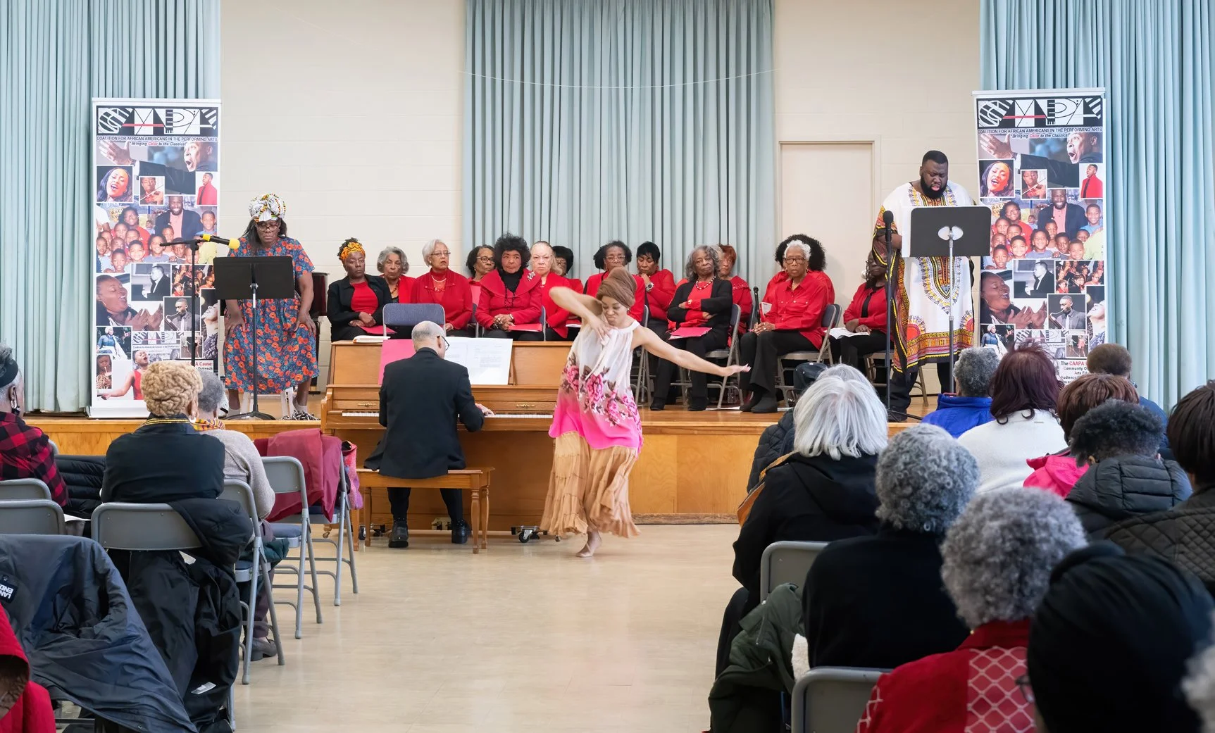 A cultural event with a dance performance in front of a choir. The choir members, mostly women dressed in red blouses, are seated on stage behind a woman who is dancing. A man is playing the piano, and two speakers stand on either side of the stage.