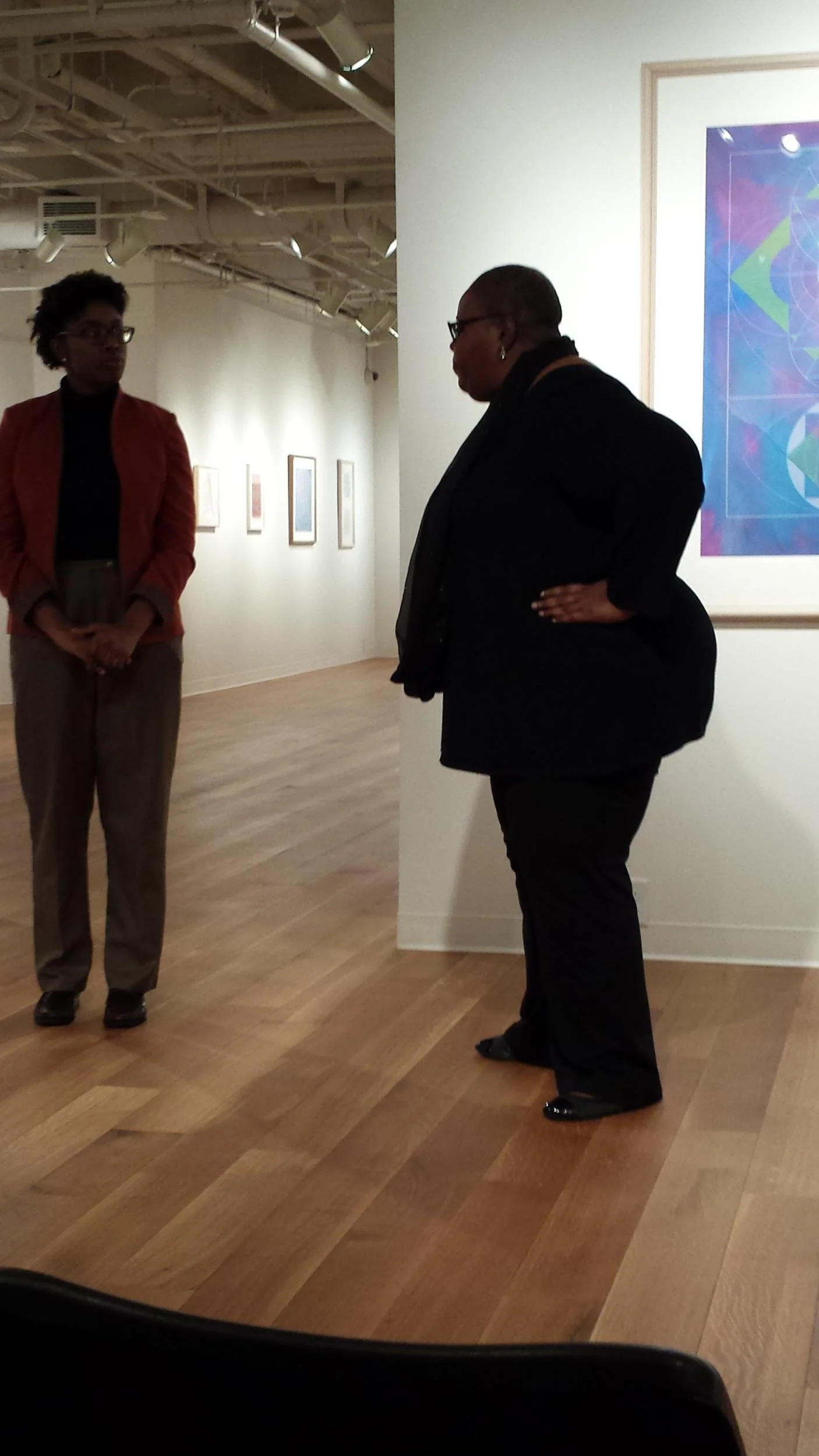 Two women are having a conversation in an art gallery. One woman wears a black outfit with glasses, and the other in a rust-colored blazer and beige trousers. Artworks are displayed on the wall in the background.