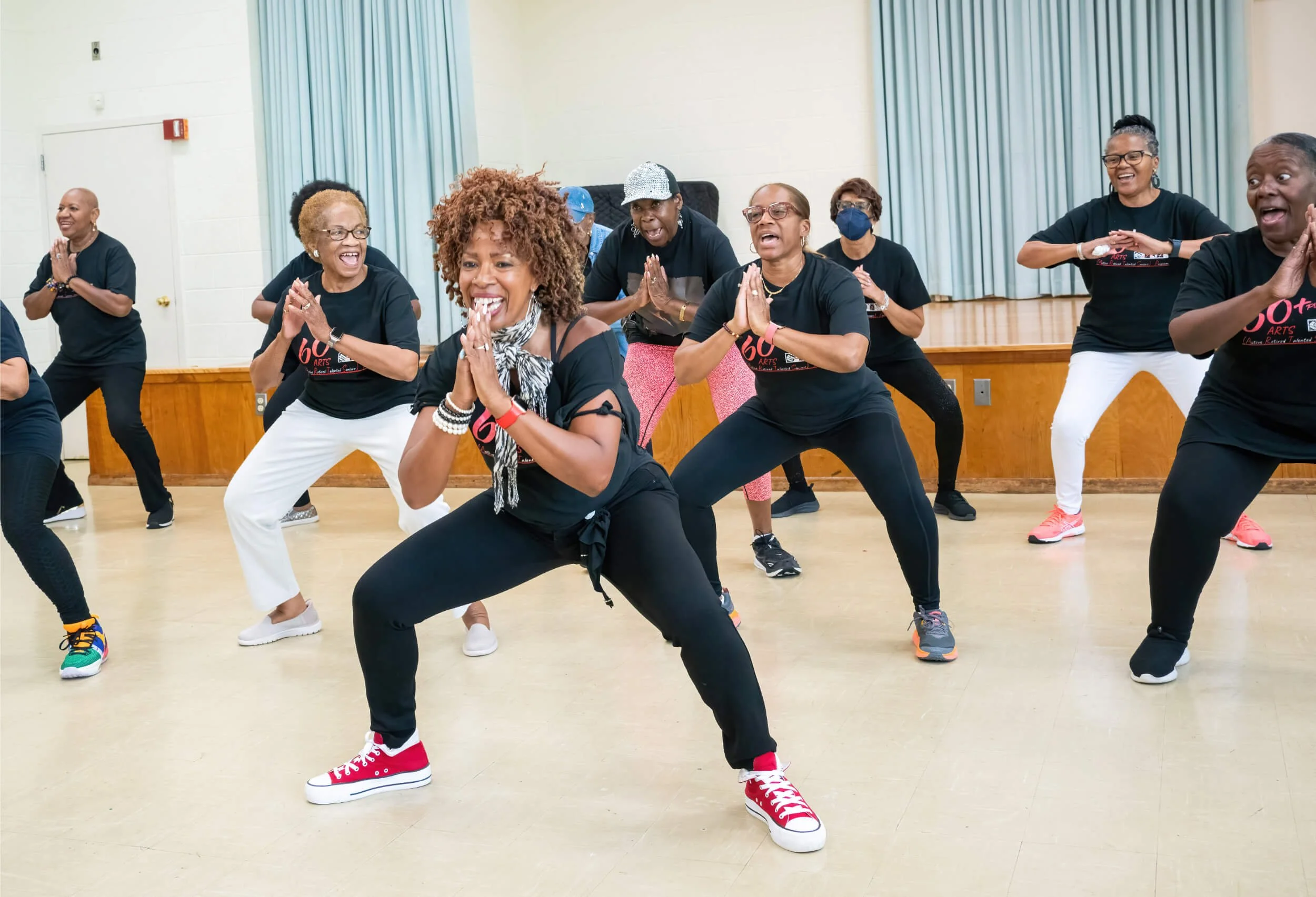 Group of women participating in a dance class in a community hall, wearing black T-shirts and colorful sneakers, enjoying and smiling during the activity.