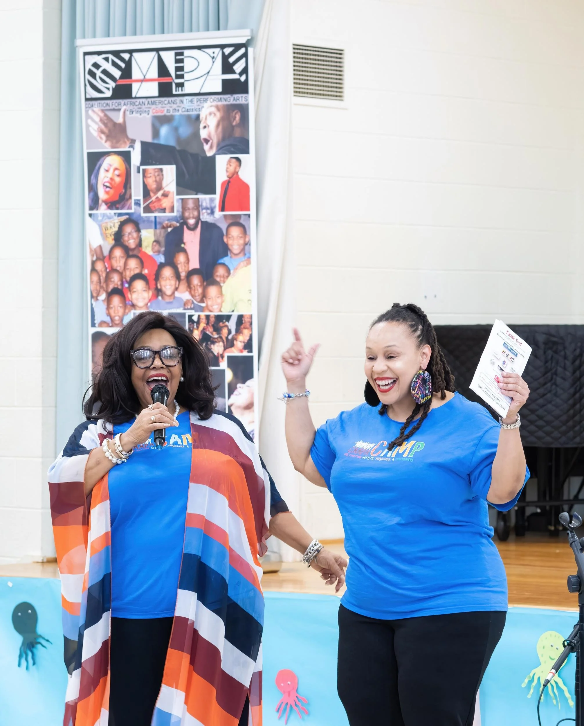 Two women on stage, one speaking into a microphone and the other smiling and raising her hand, with a poster of diverse African American children and adults in the background.