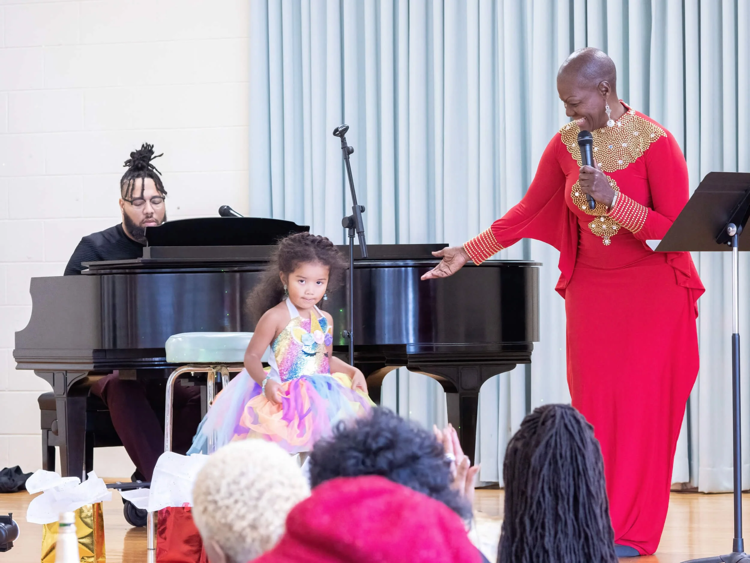 A woman in a red dress with gold embellishments is holding a microphone and gesturing towards a young girl in a colorful rainbow dress sitting on a chair next to a black grand piano. A man is playing the piano in the background, and there are people
