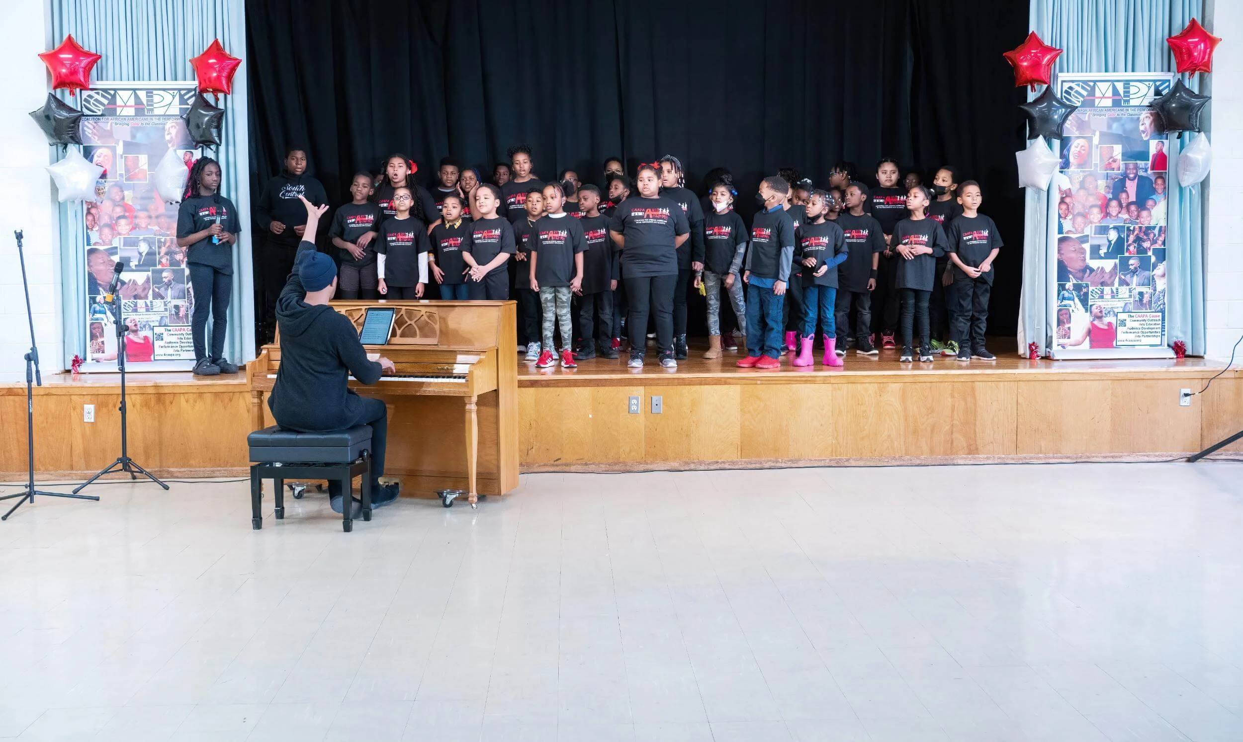 Children and adults on stage singing or performing, with a pianist playing at a keyboard in front of them. The stage is decorated with black curtains, red, white, and silver star balloons, and posters on either side.