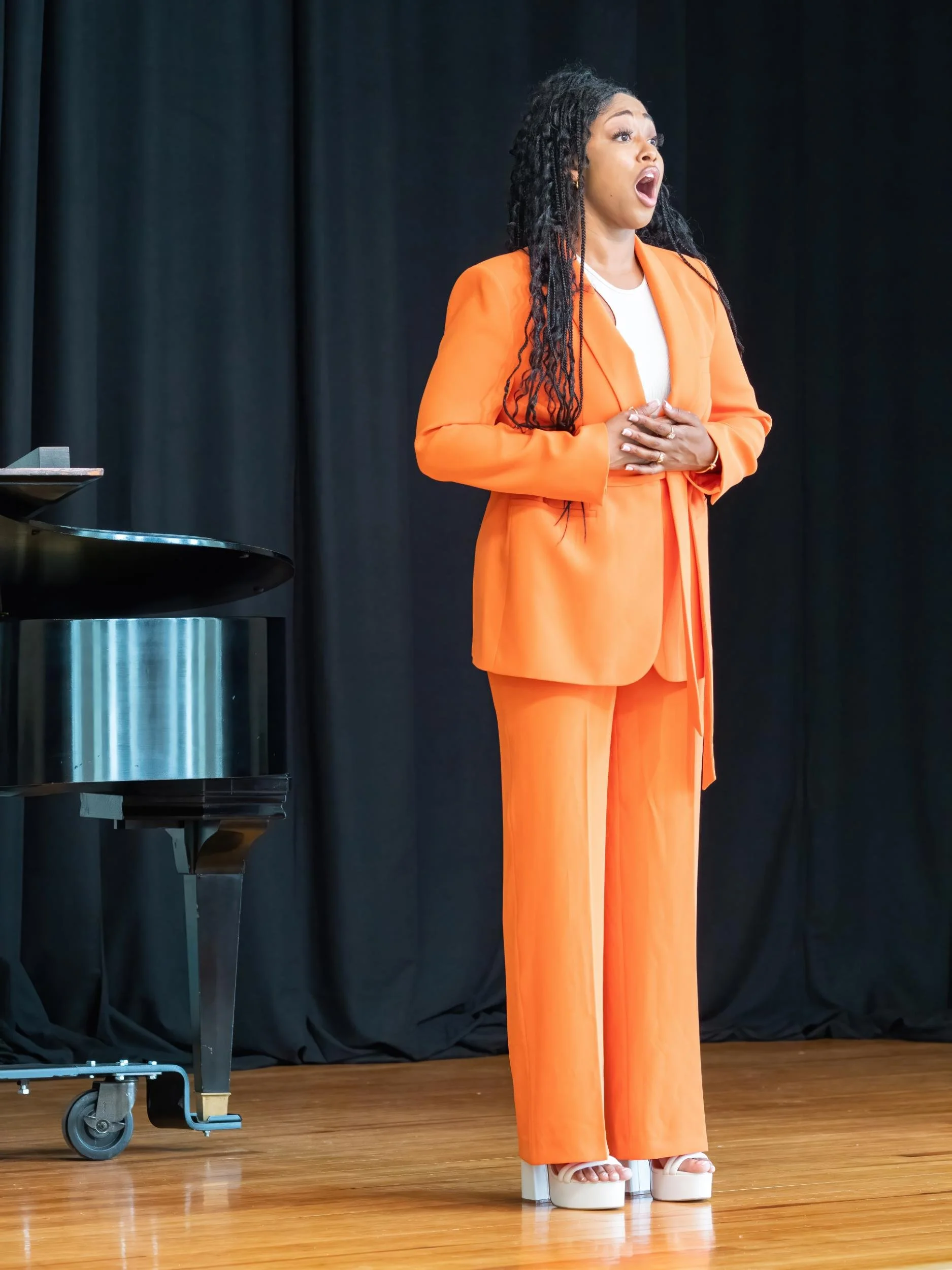 A woman wearing an orange suit and high heels, standing on a wooden stage with a black curtain in the background, appears to be singing or speaking passionately.