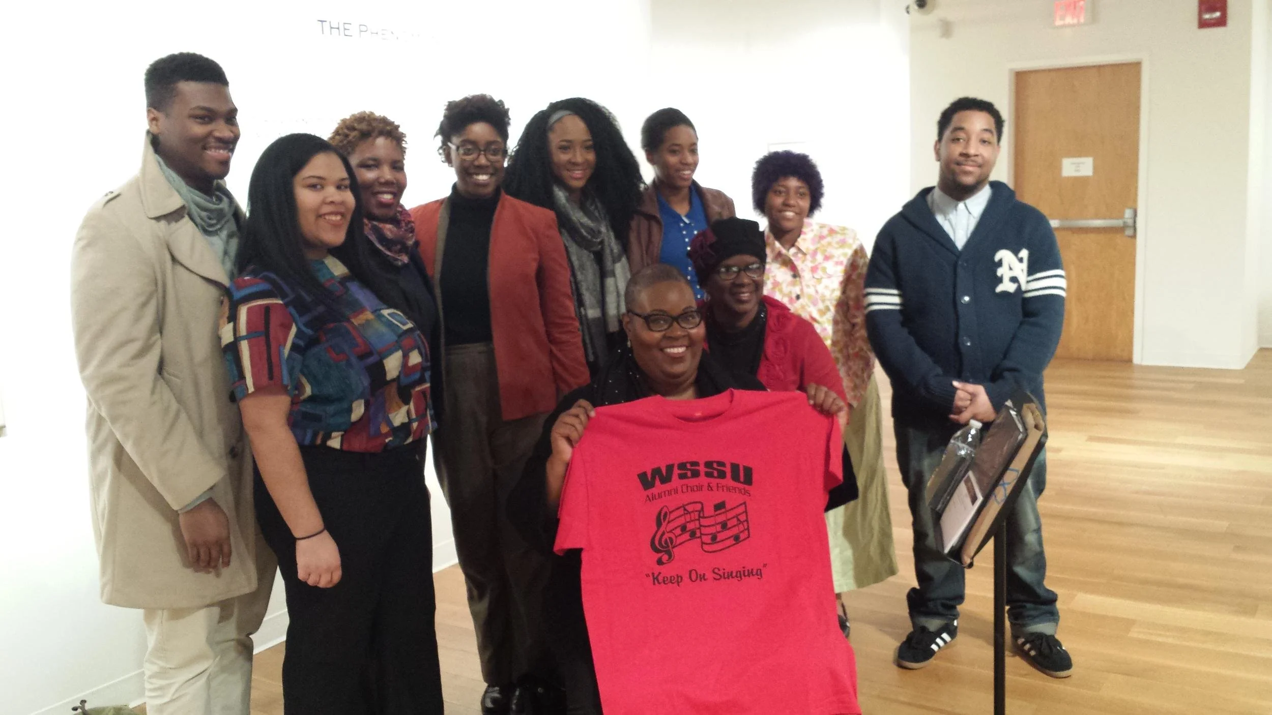 Group of ten diverse people, women and men, smiling indoors with a plain white wall and wooden floor, holding a red T-shirt with 'WSSU Alumni Choir & Friends' and 'Keep On Singing' printed on it.