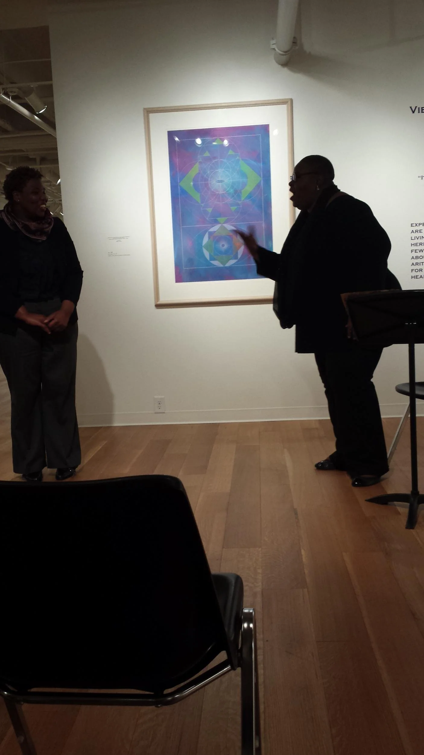 Two women are standing and listening to a man who is explaining or discussing artwork in an art gallery. The artwork behind them is an abstract, geometric, and colorful piece.