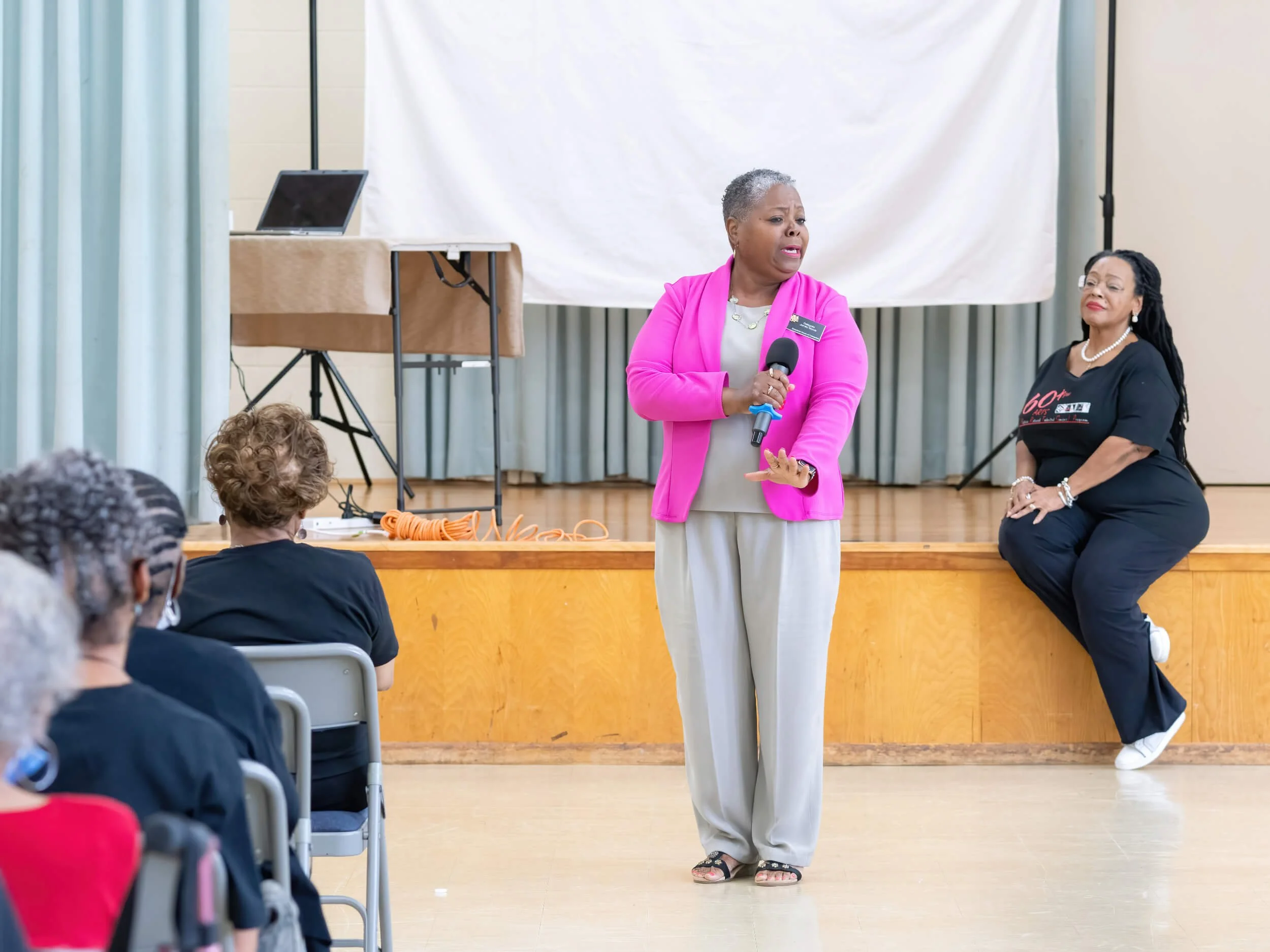 A woman in a pink blazer speaking into a microphone in front of an audience, with another woman sitting on the stage behind her; the audience includes elderly women.