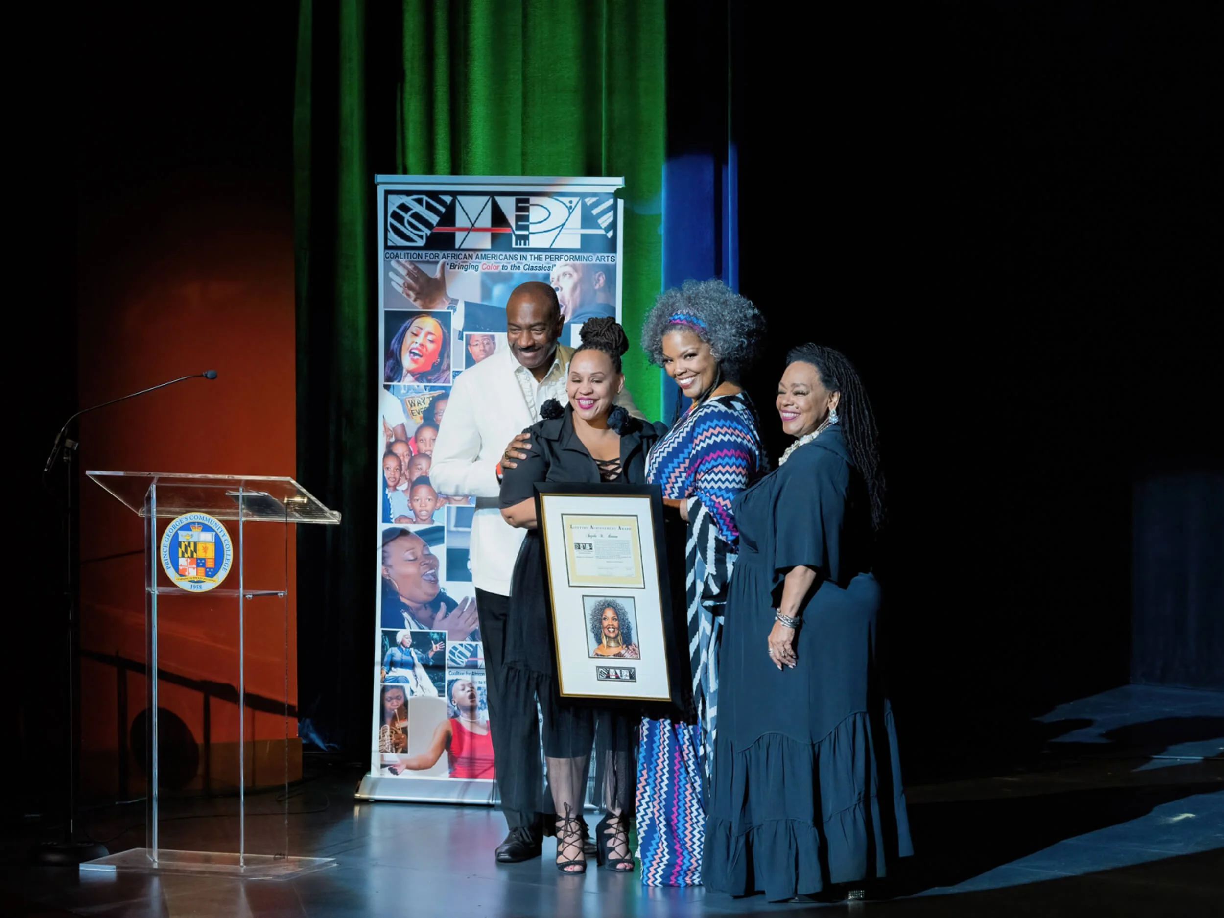 Four people standing on a stage, holding a framed certificate and photo, at an event celebrating African Americans in the performing arts. A banner with images and text is behind them, and there's a transparent lectern with a seal on it to the left.