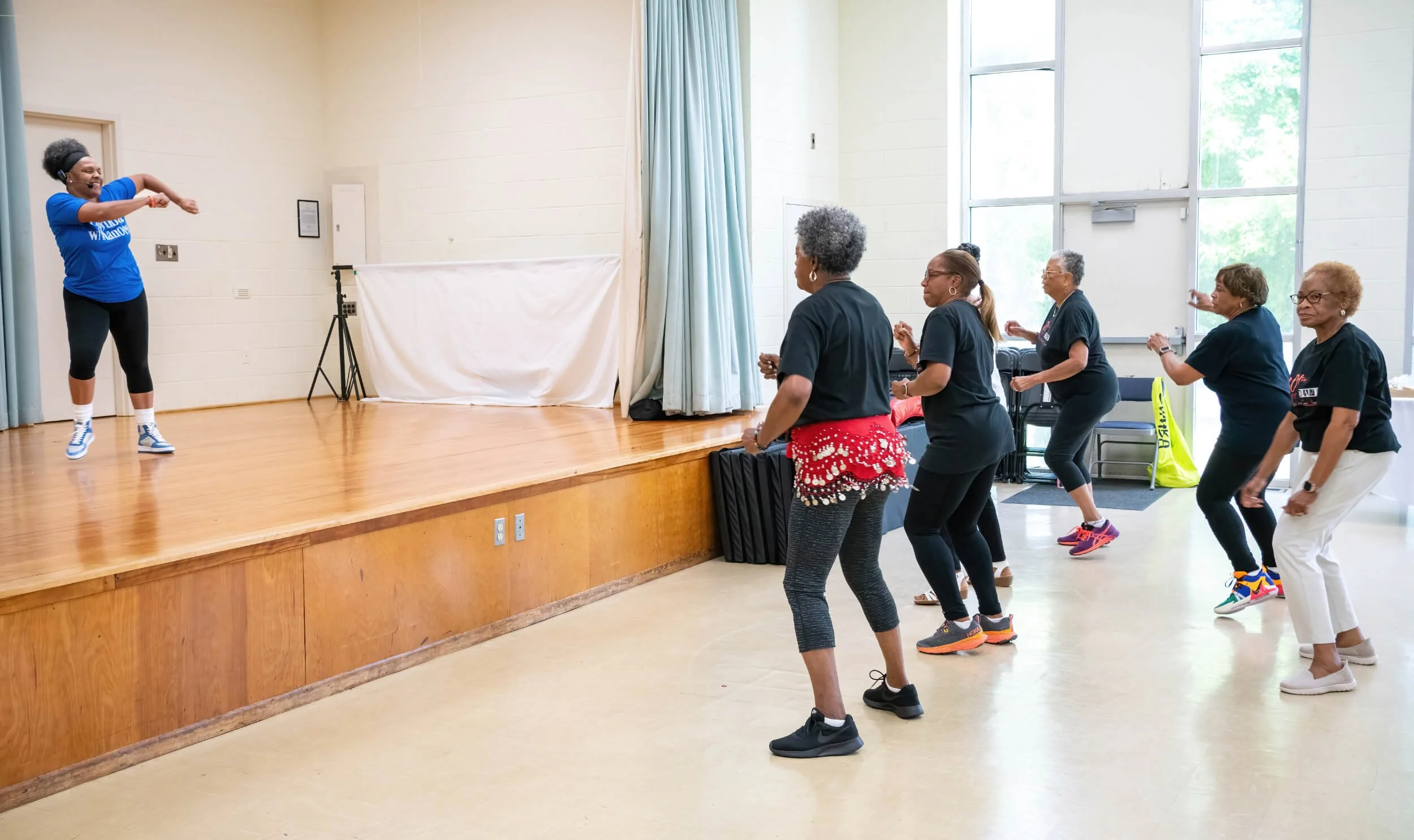 A group of women participating in a dance class or exercise session led by an instructor on stage in a spacious room with large windows, hardwood floor, and light-colored walls.