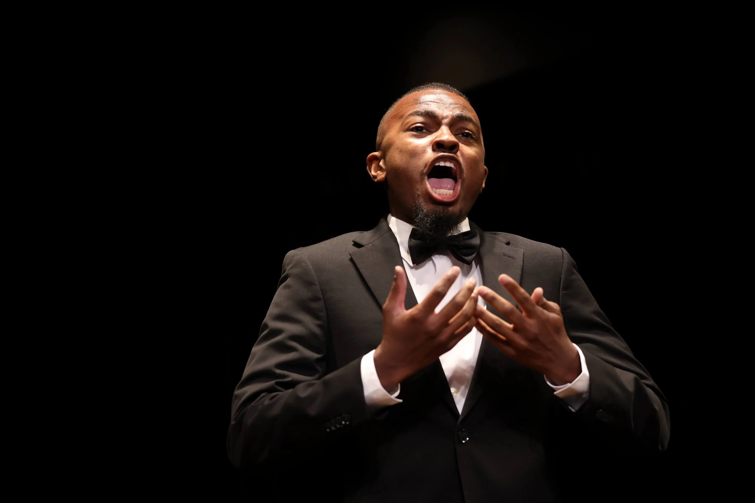 A man in a tuxedo and bow tie passionately singing or performing on stage with a black background.