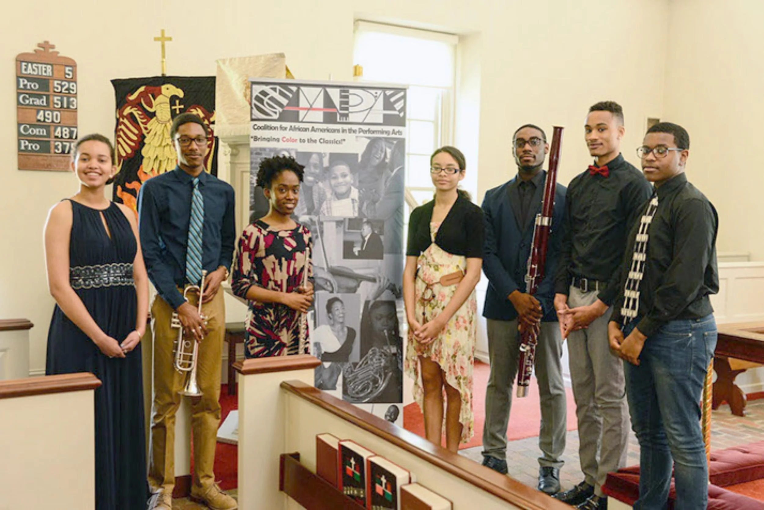 Group of eight young people standing inside a church, dressed in formal clothes, with musical instruments and a banner. The church wall behind has a hymn board and a decoration with a cross and eagle emblem.