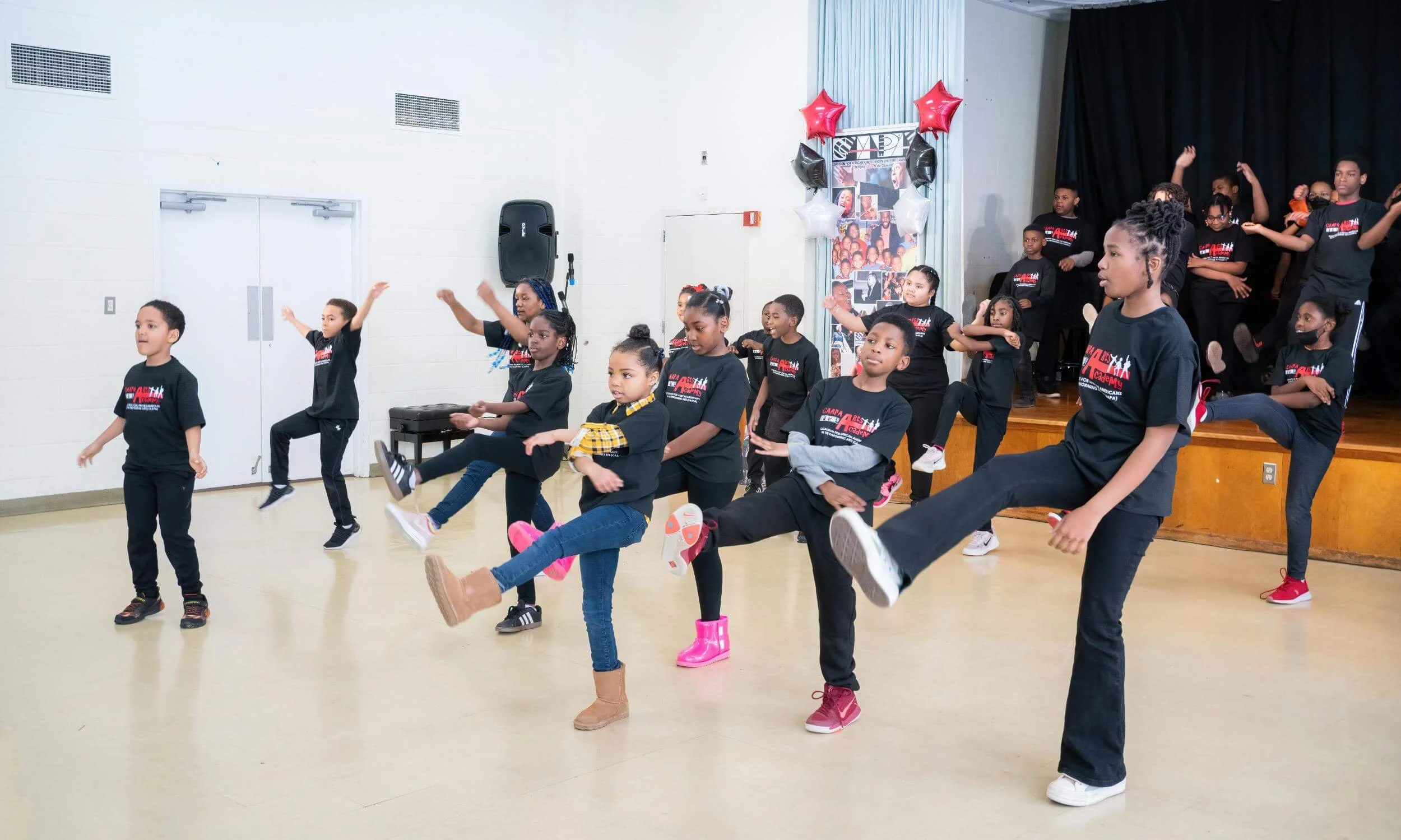 Group of children performing a dance or exercise in a school auditorium with red, white, and black balloons and football themed decorations.