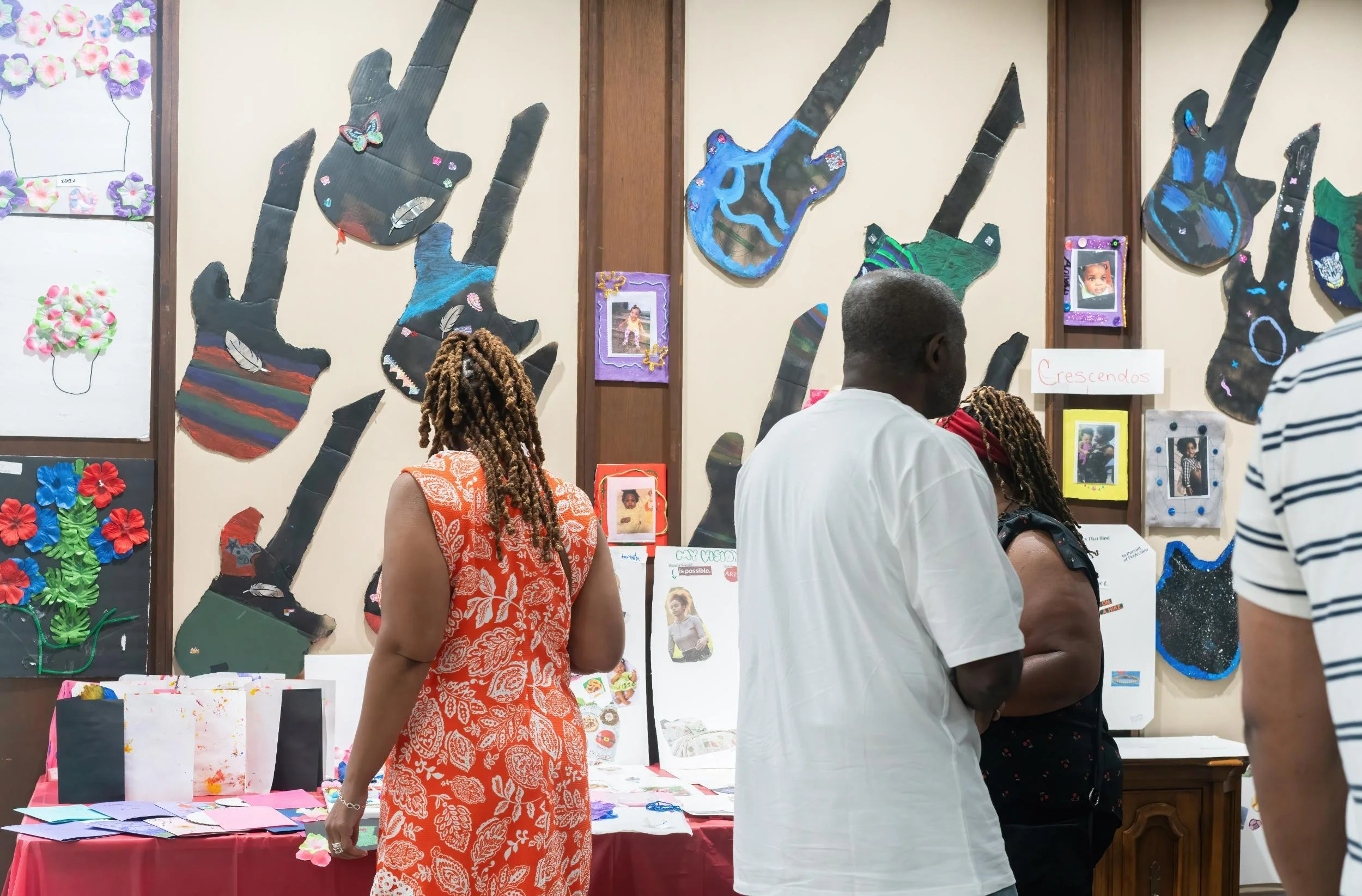 People viewing artwork and crafts displayed on a wall and table at an art exhibition, with handmade decorated guitars and framed pictures.