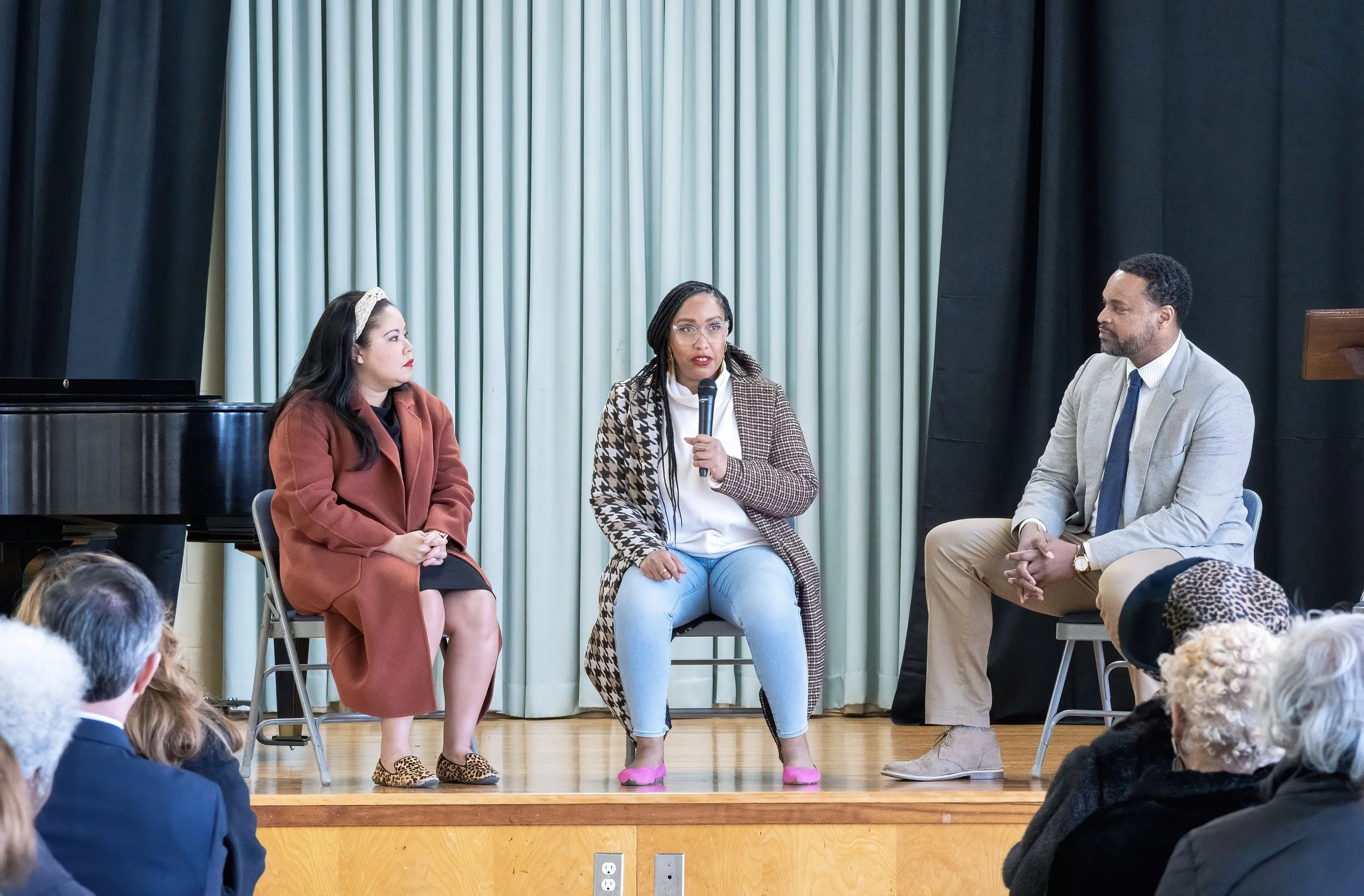 Three people on stage speaking at an event, with an audience listening. One woman in the middle is holding a microphone, wearing glasses and a brown and white patterned coat. To her left, a woman in a reddish-brown coat and leopard print shoes, and t