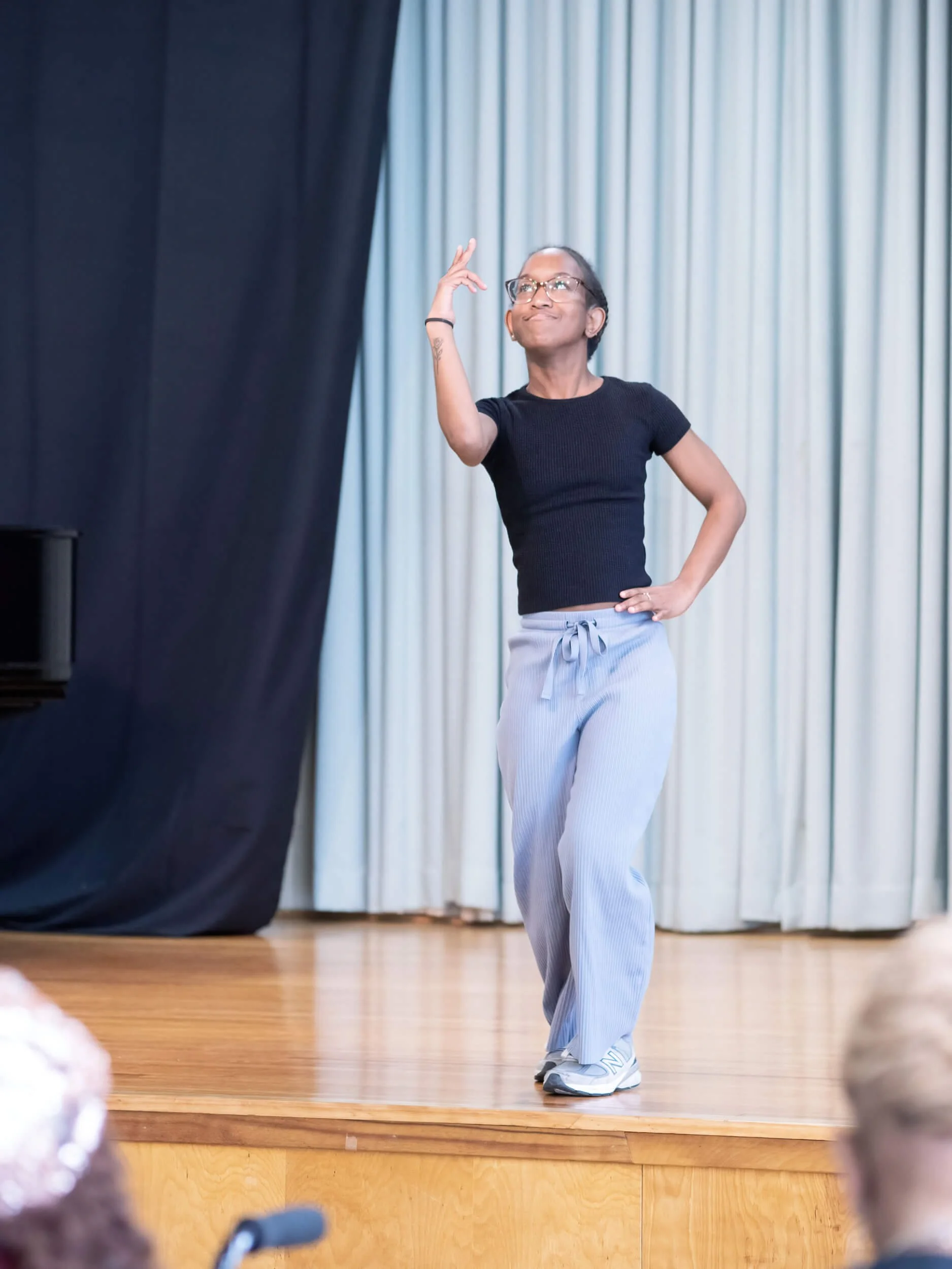 A young woman with glasses and a black t-shirt performing a dance or theatrical pose on stage with light-colored curtains in the background.