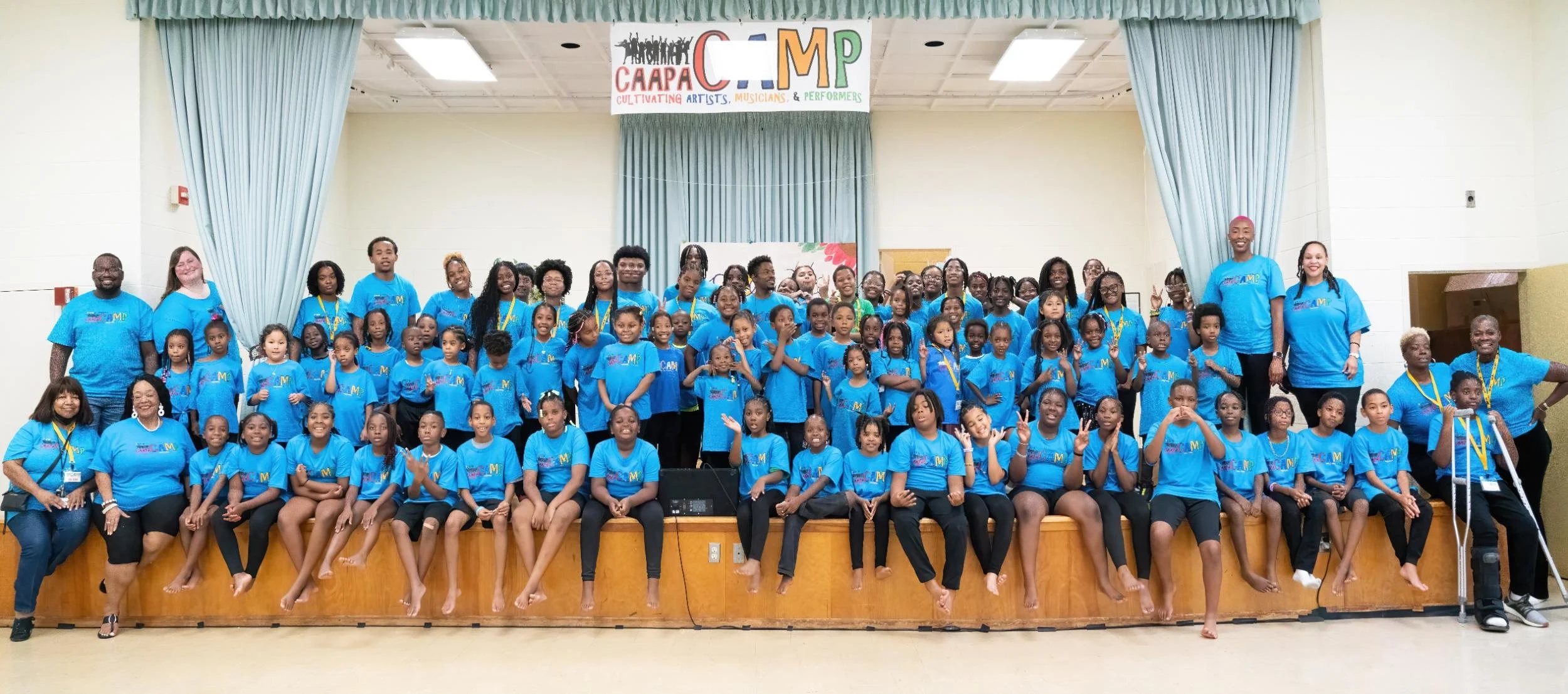 A large group of children and adults on a stage for a group photo at an event called CAAPA CAMP, with a banner above that reads 'Cultivating Artists, Musicians, & Performers.' All are wearing matching blue T-shirts with the C A A P M logo.