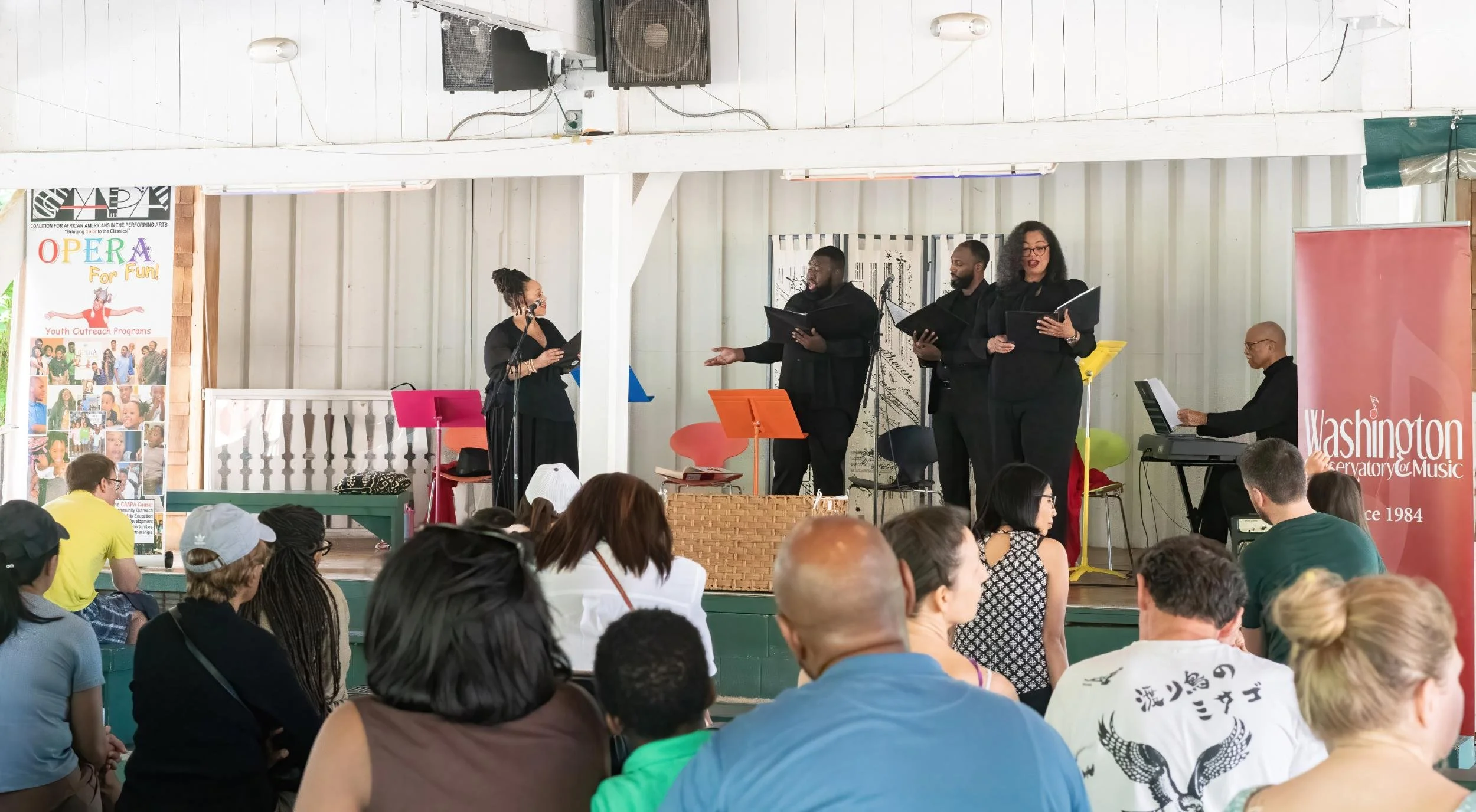 A choir performs on stage at a community event, with a woman singing into a microphone, four people holding songbooks, and a man playing keyboard, while an audience watches nearby.