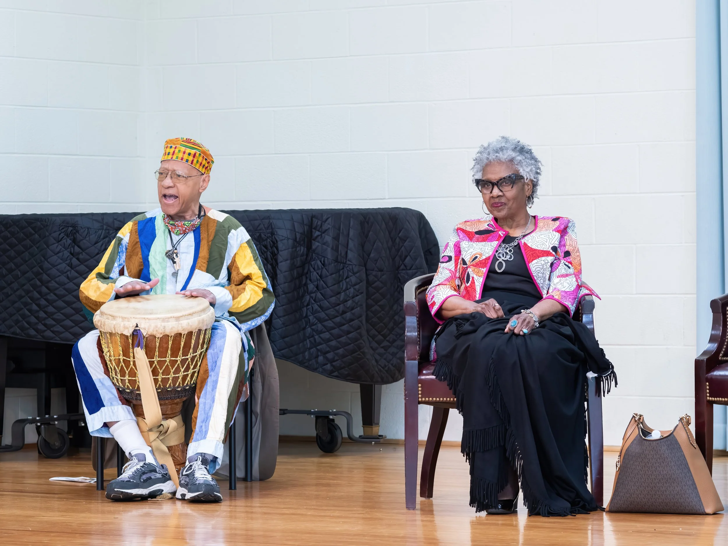 An elderly man playing a drum and singing, sitting on a chair. An elderly woman with gray curly hair, wearing glasses and a colorful blazer, sits with her arms crossed, listening intently. Both are seated in a room with a white brick wall and wooden