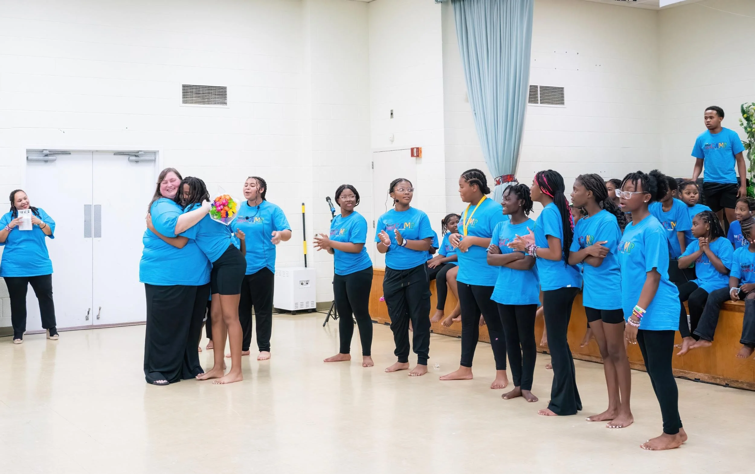 A group of young girls wearing blue t-shirts, some with medals, standing on a stage in a school auditorium, clapping and smiling as they watch a woman hug a younger girl. Other girls are seated on the stage, some smiling and observing the scene.