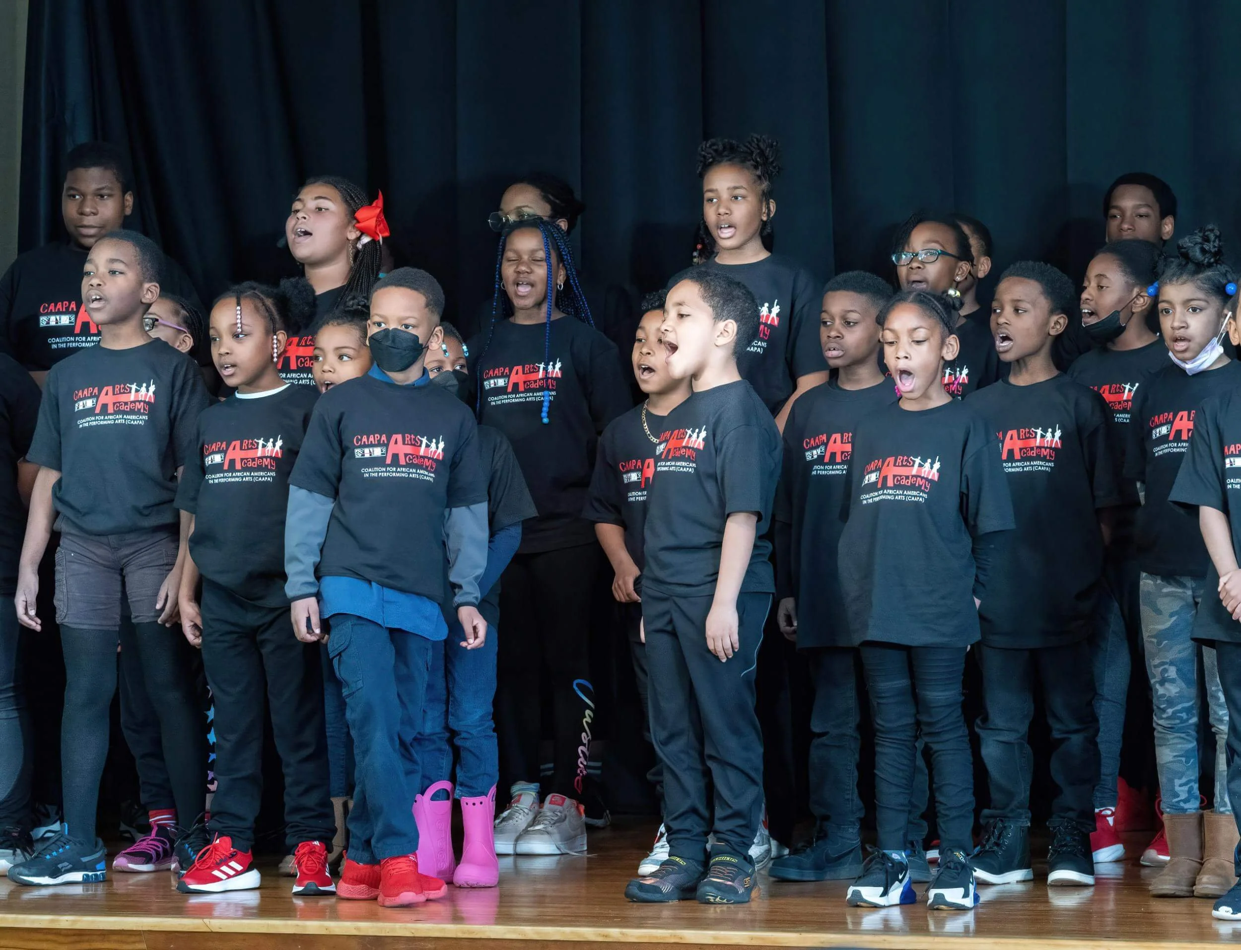 Children singing on stage wearing black T-shirts with red and white text and graphics, a dark curtain backdrop.