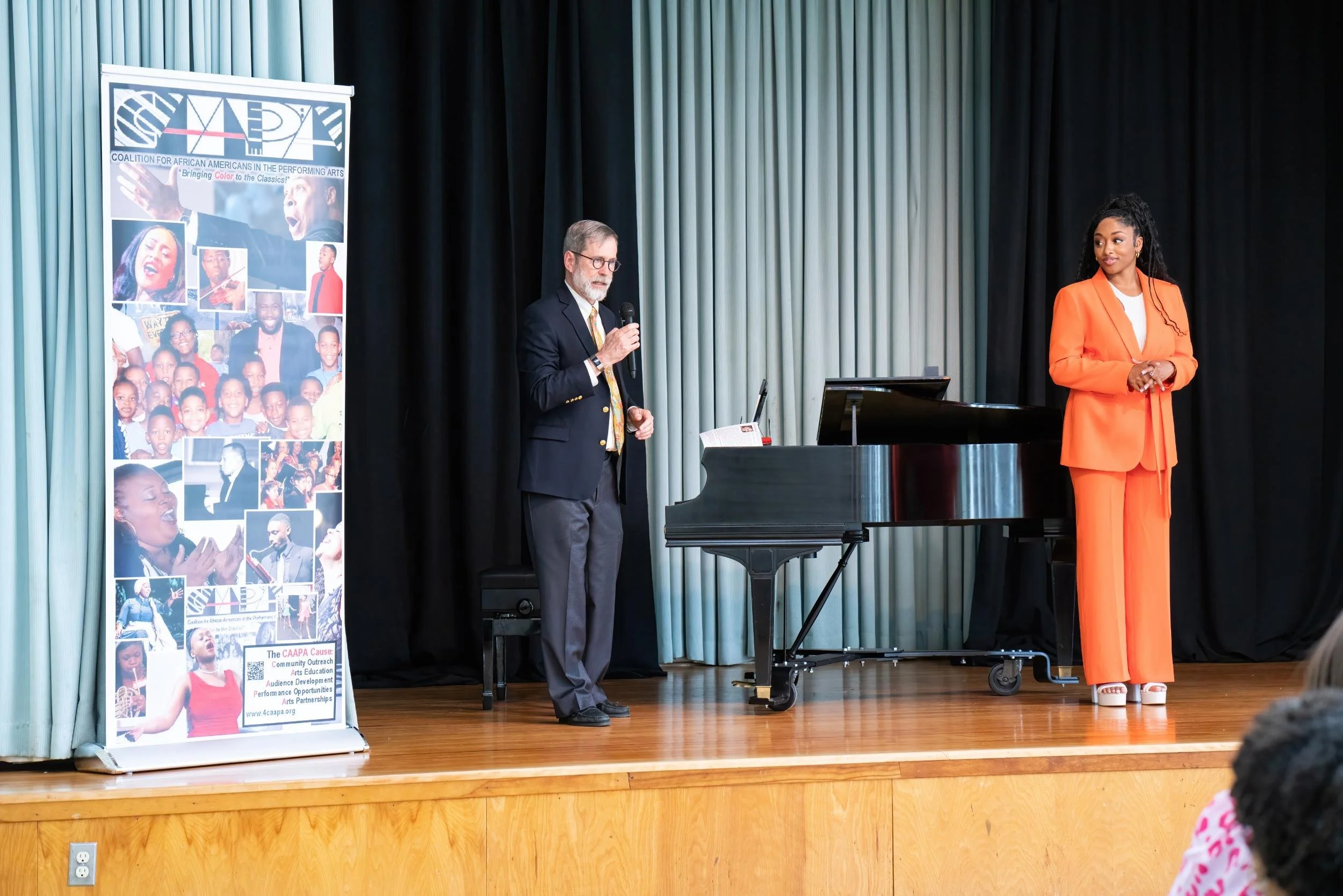 Two individuals on stage, one man in a dark suit holding a microphone, and a woman in an orange suit standing beside a black grand piano. Behind them are black and light blue curtains, and a tall banner with photos of people and text about the Coalit