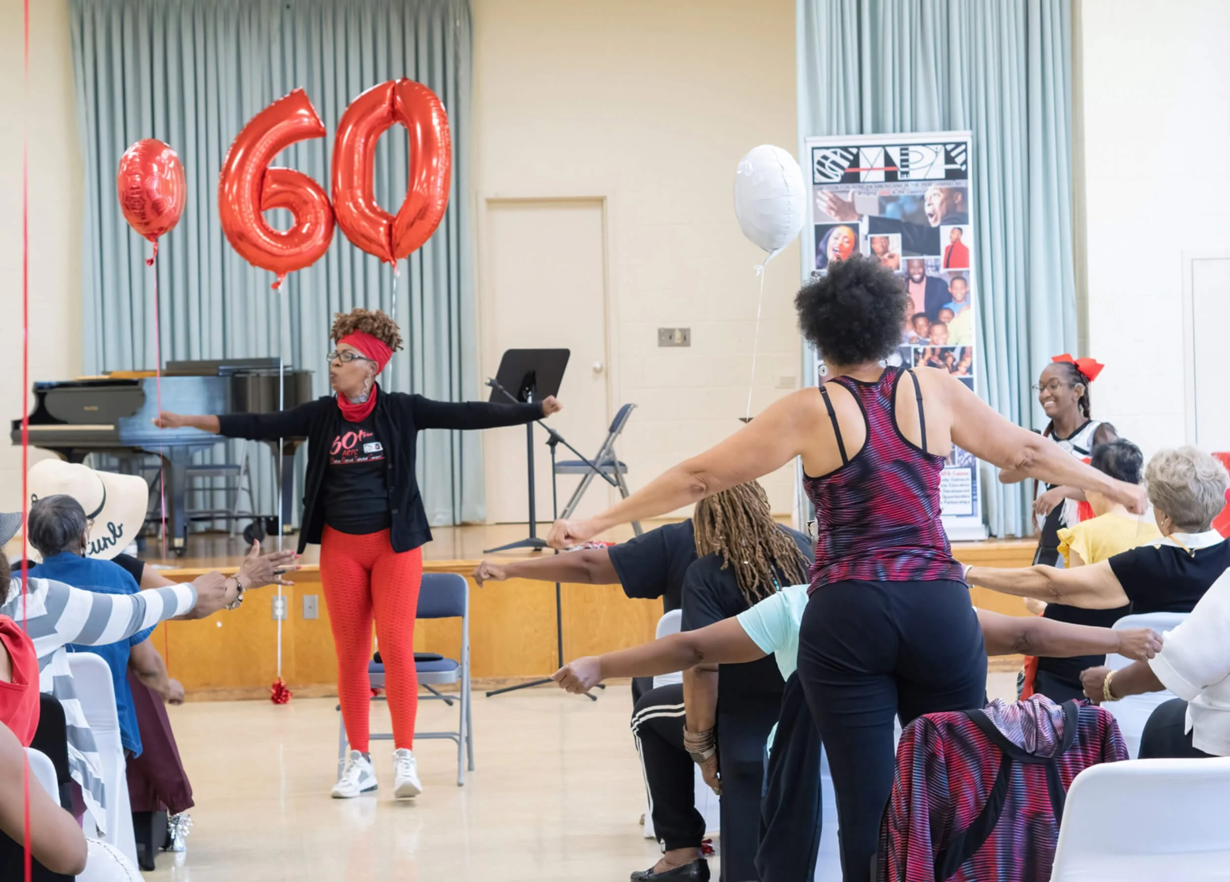 Group of people participating in a dance or exercise class in celebration of a 60th anniversary, with red and white balloons, and a woman leading the class.
