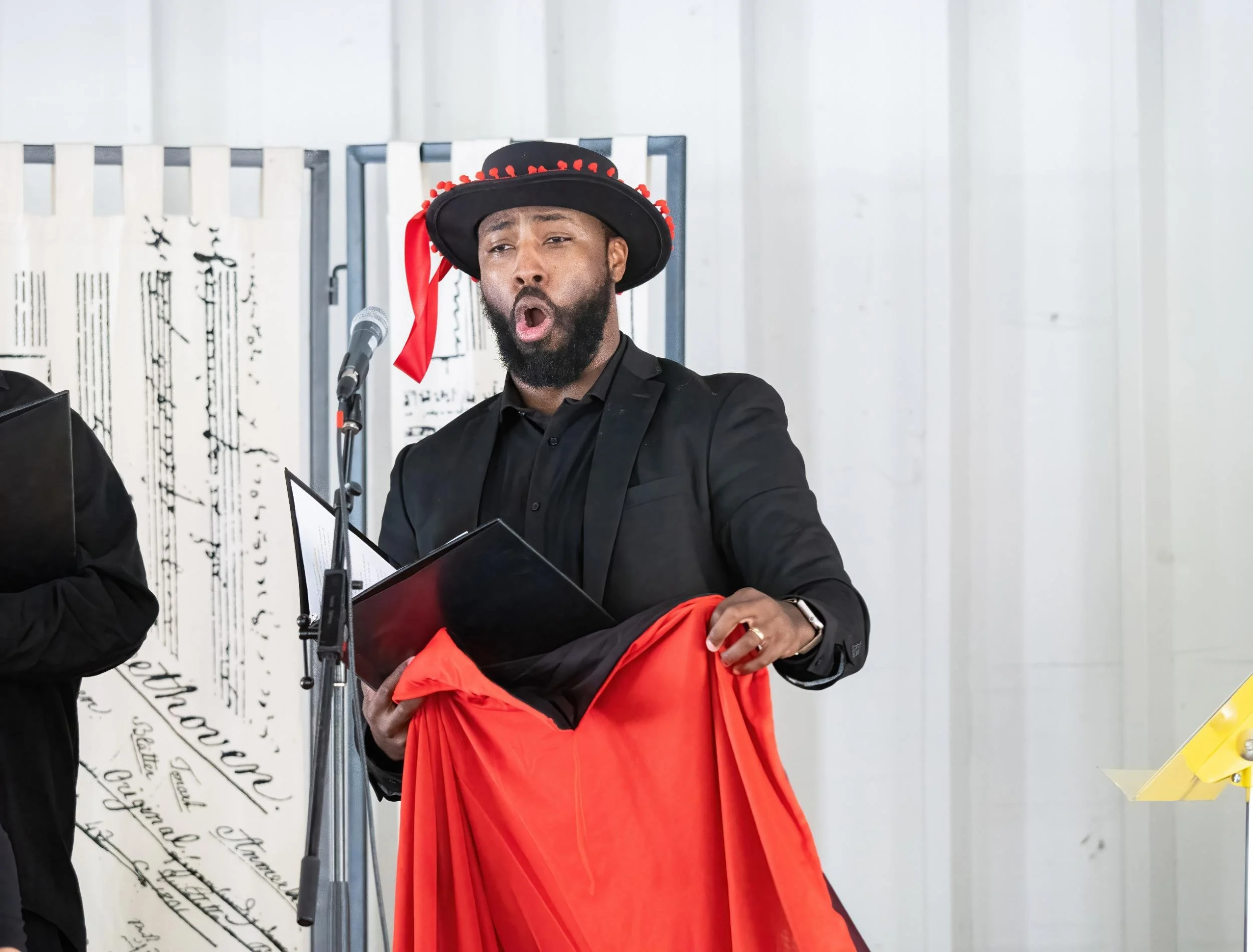 A man singing into a microphone, wearing a black hat decorated with red accents, in a formal black suit, holding a red cloth and black folder, in a setting with white panel walls and music sheets in the background.