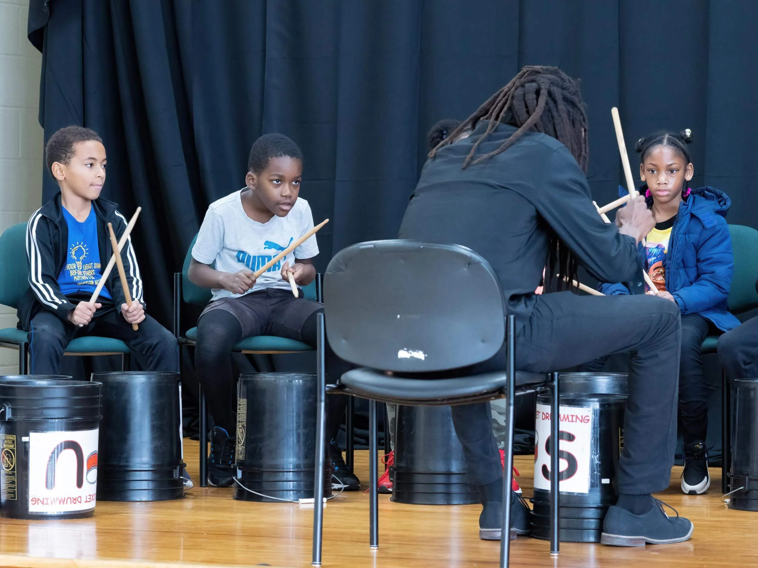 Children sitting on chairs holding drumsticks during a drumming lesson or performance, with a person demonstrating in front of them.