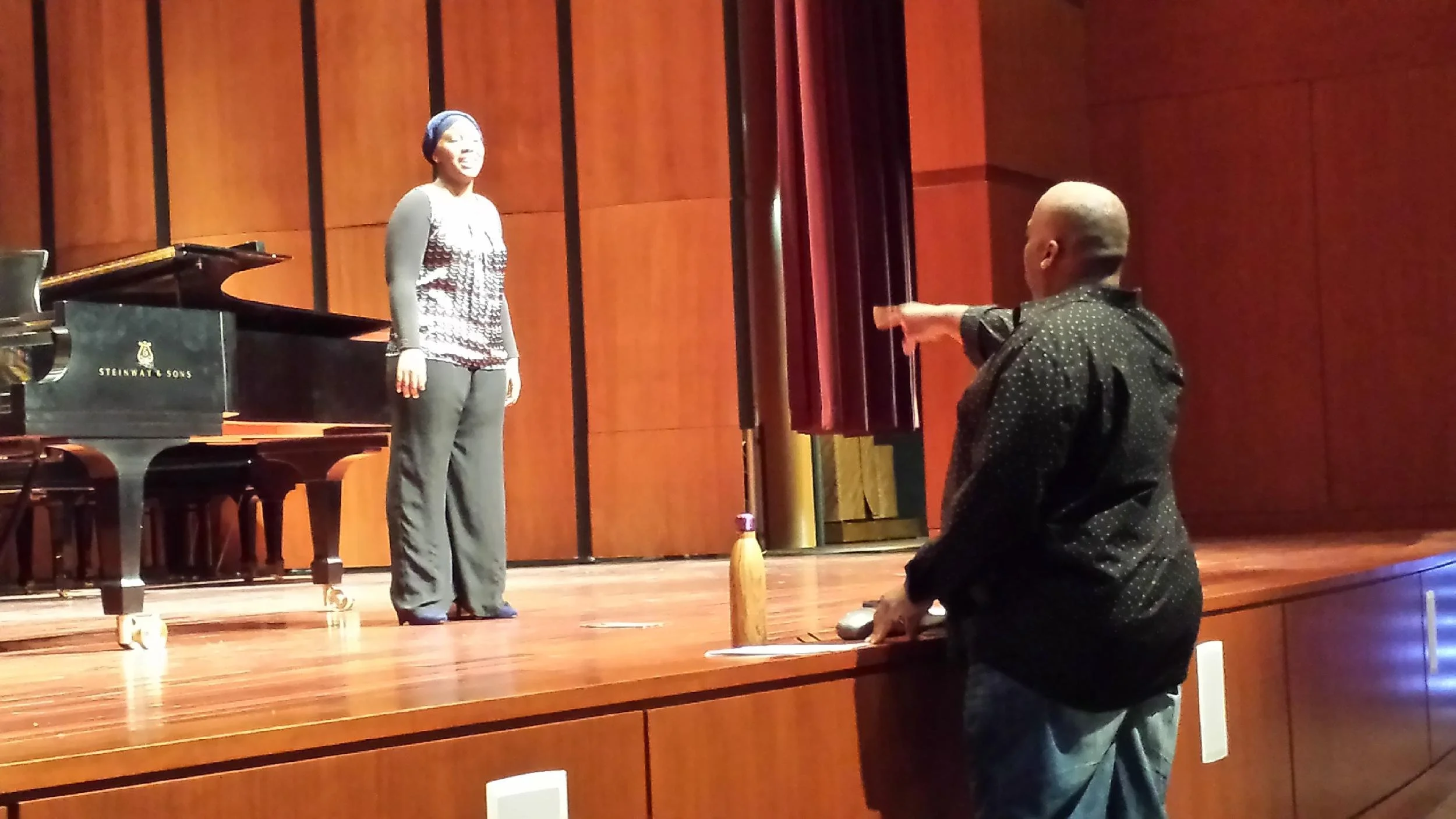 A person on stage pointing at a woman standing near a piano, with a wooden wall background.