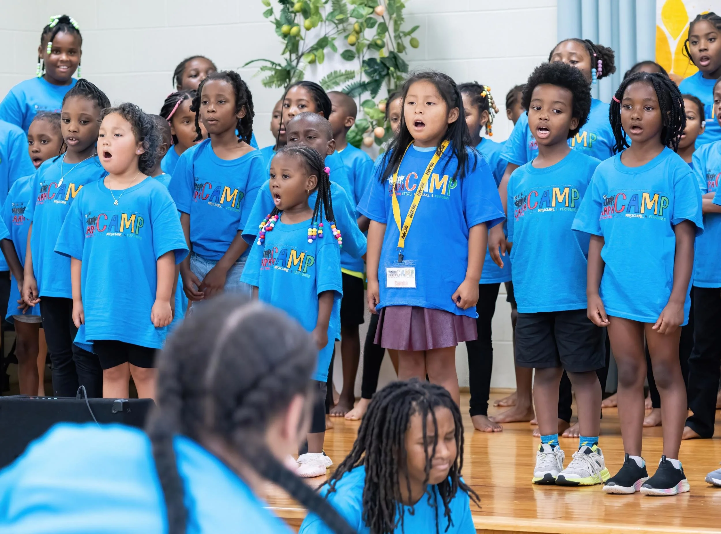 A group of children singing on stage at a camp, wearing blue T-shirts that say 'Camp'.