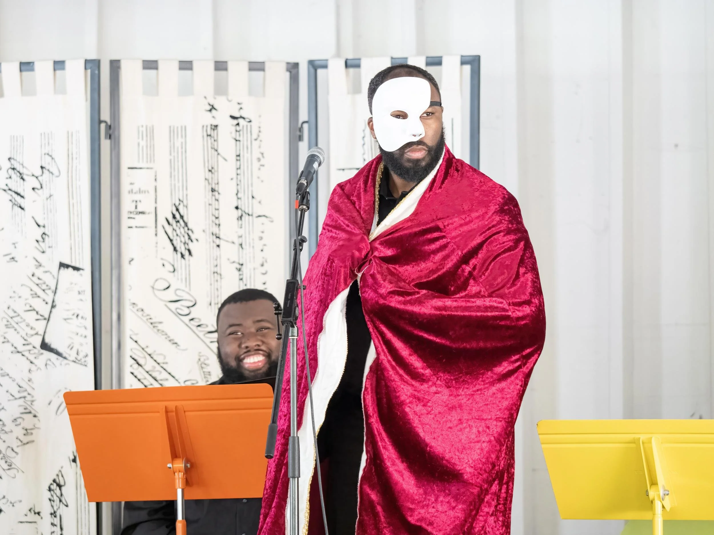 A man in a gold and red costume with a white mask over half his face stands with a serious expression in front of a microphone. Behind him, a smiling musician plays a keyboard, with sheet music on stands. The background has a white decorative folding