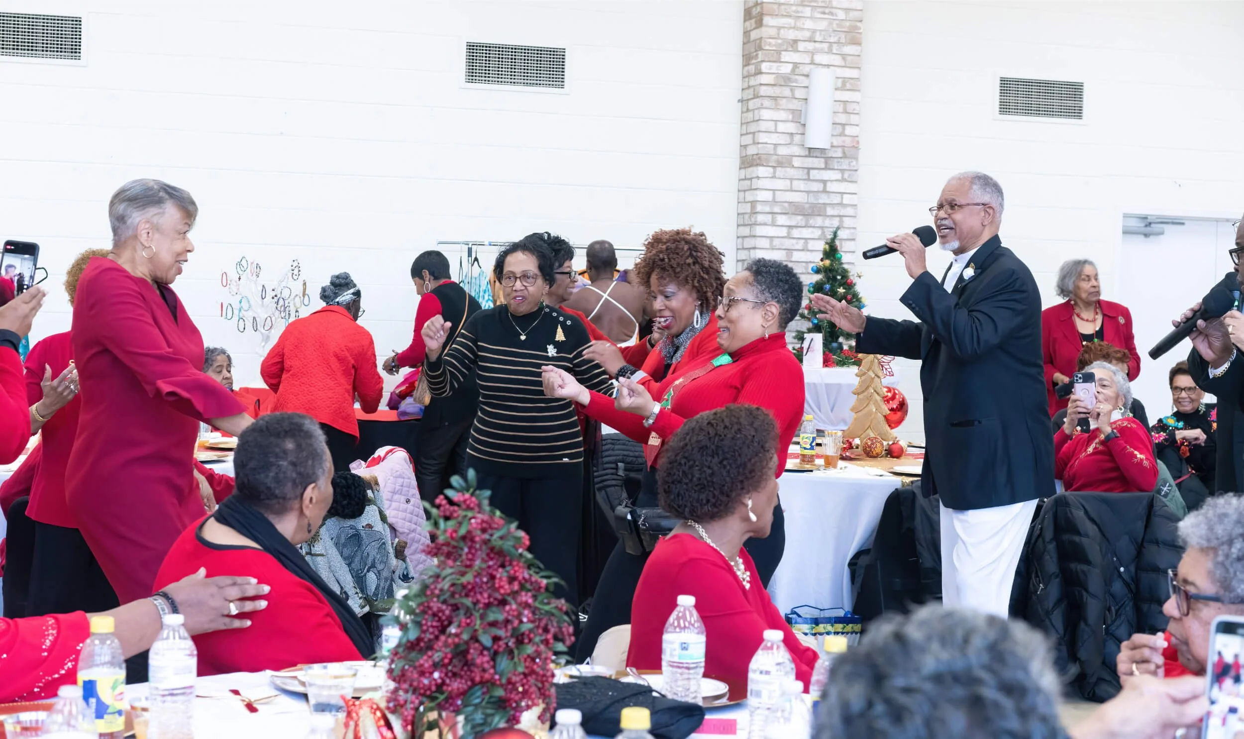 Group of people at a holiday celebration, some sitting and others dancing and singing, decorated with Christmas trees and ornaments.