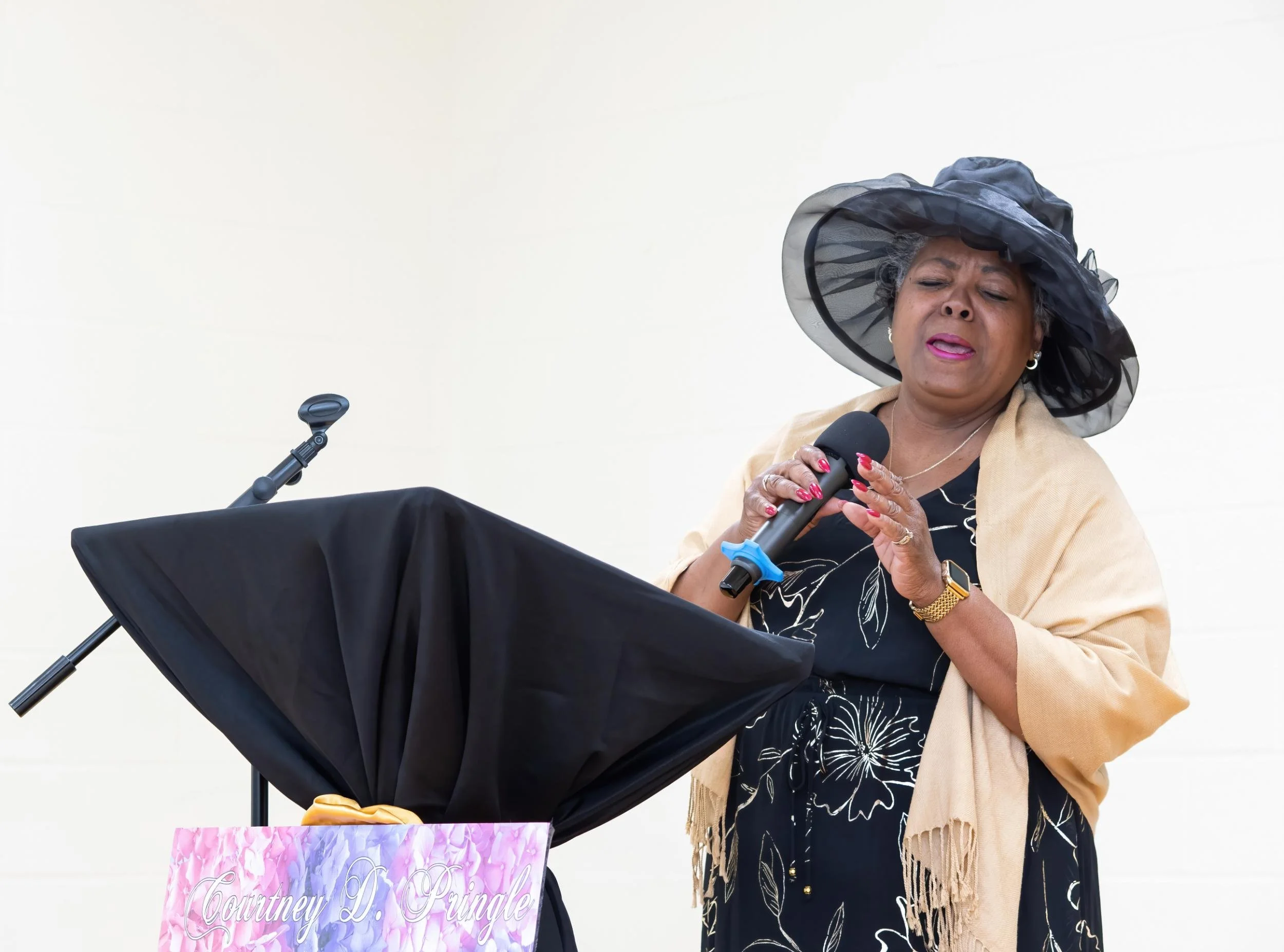 A woman wearing a black hat, beige shawl, and a black dress with white floral patterns, sings into a microphone at a memorial service, standing behind a podium with a floral sign that reads 'Courtney D. Pingle'.