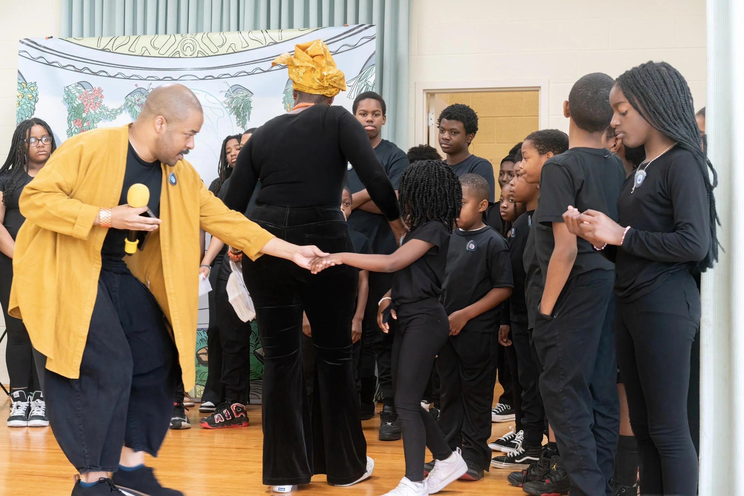 A man with a yellow jacket shaking hands with a young girl in black clothing during a group gathering or event, with multiple children and adults present in the background.
