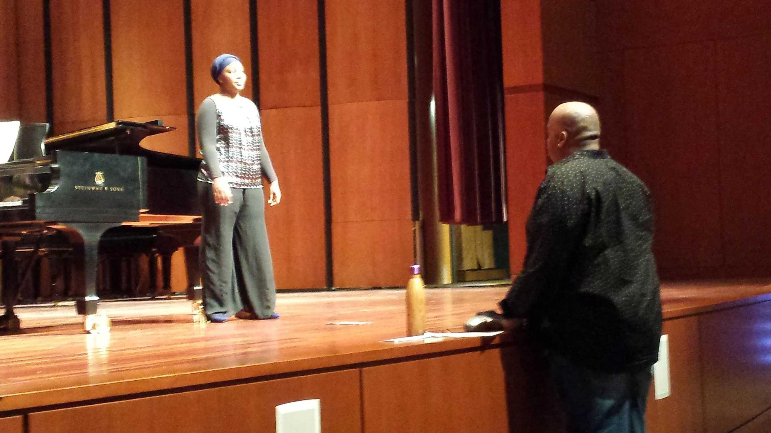 A woman standing on a stage near a grand piano, wearing a headscarf and colorful sweater, with a man standing at the edge of the stage, facing her.