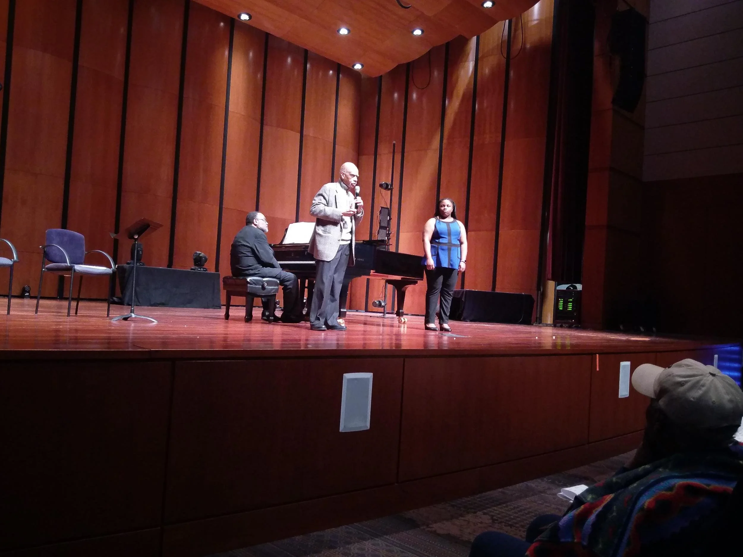 A person is speaking into a microphone on stage with a piano behind him, while another person stands nearby. An audience member is visible in the foreground.