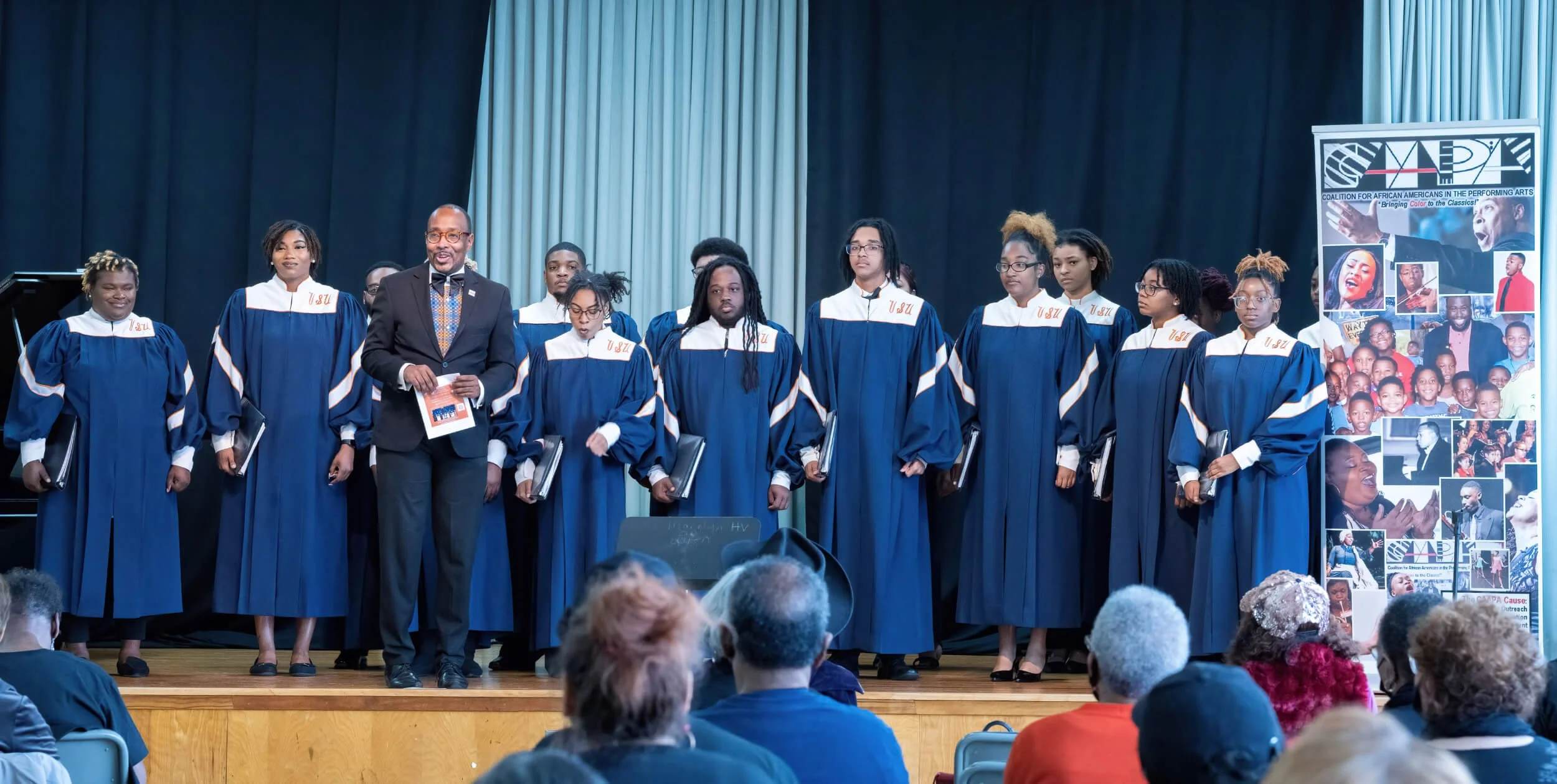 Group of graduates in blue caps and gowns standing on stage with an audience watching. A man in a black suit holding a certificate stands among them, with a banner displaying photos and text about African Americans in the performing arts.