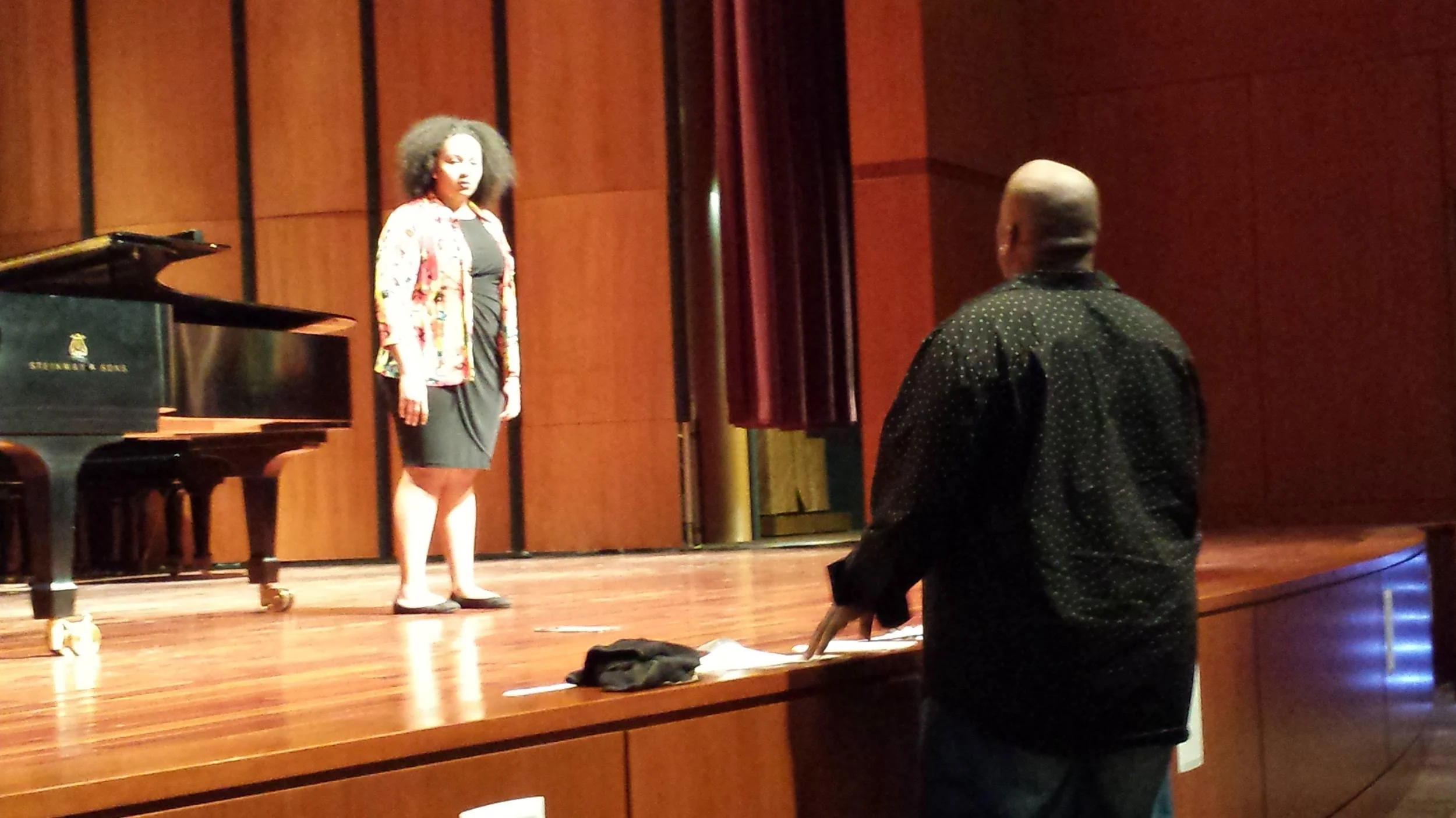 A woman singing on a stage with a grand piano nearby, and a man standing in front of her, facing the stage.