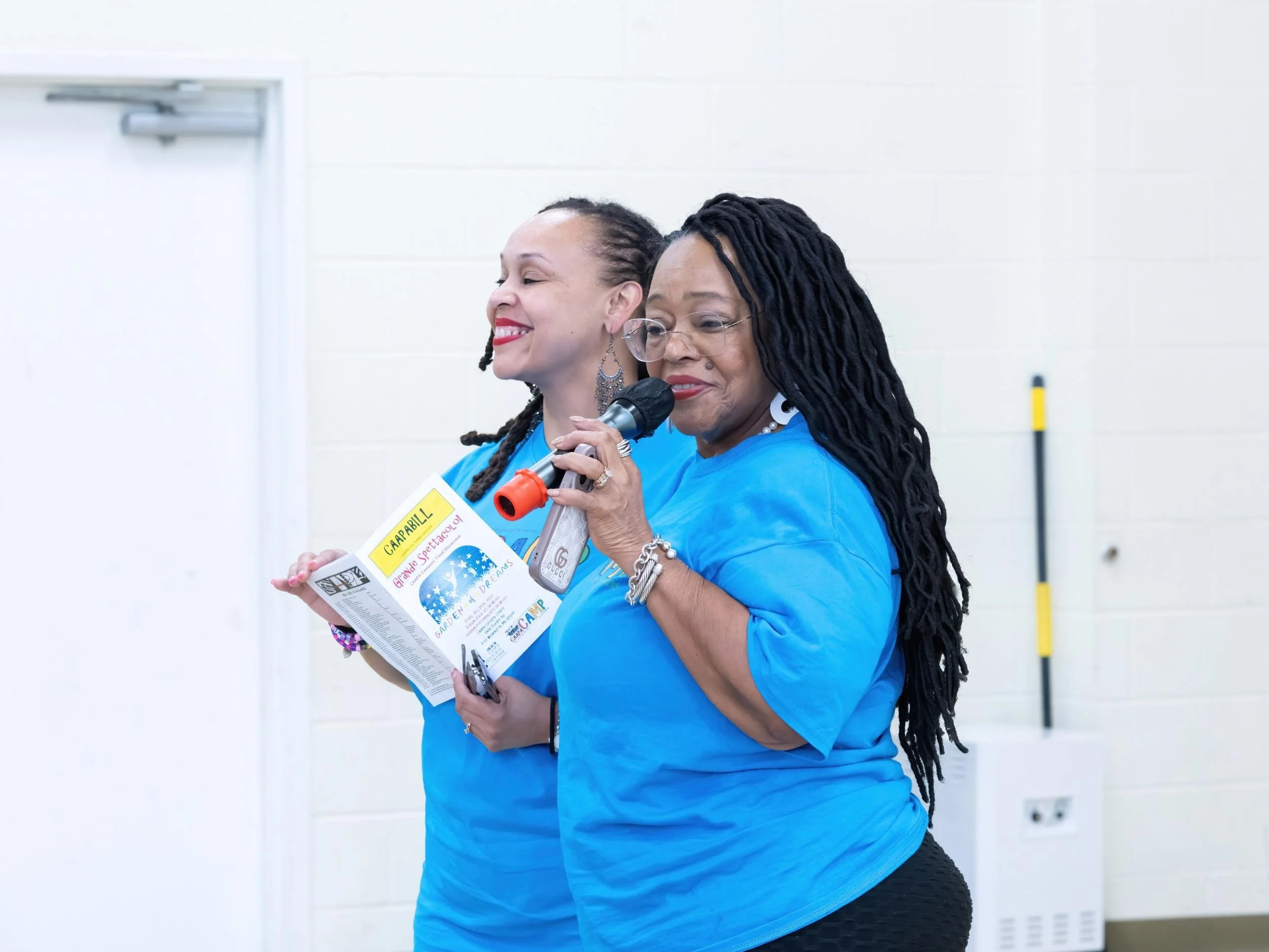 Two women wearing blue shirts, smiling, and standing indoors. One woman is holding a microphone and a pamphlet, with the other woman standing behind her.
