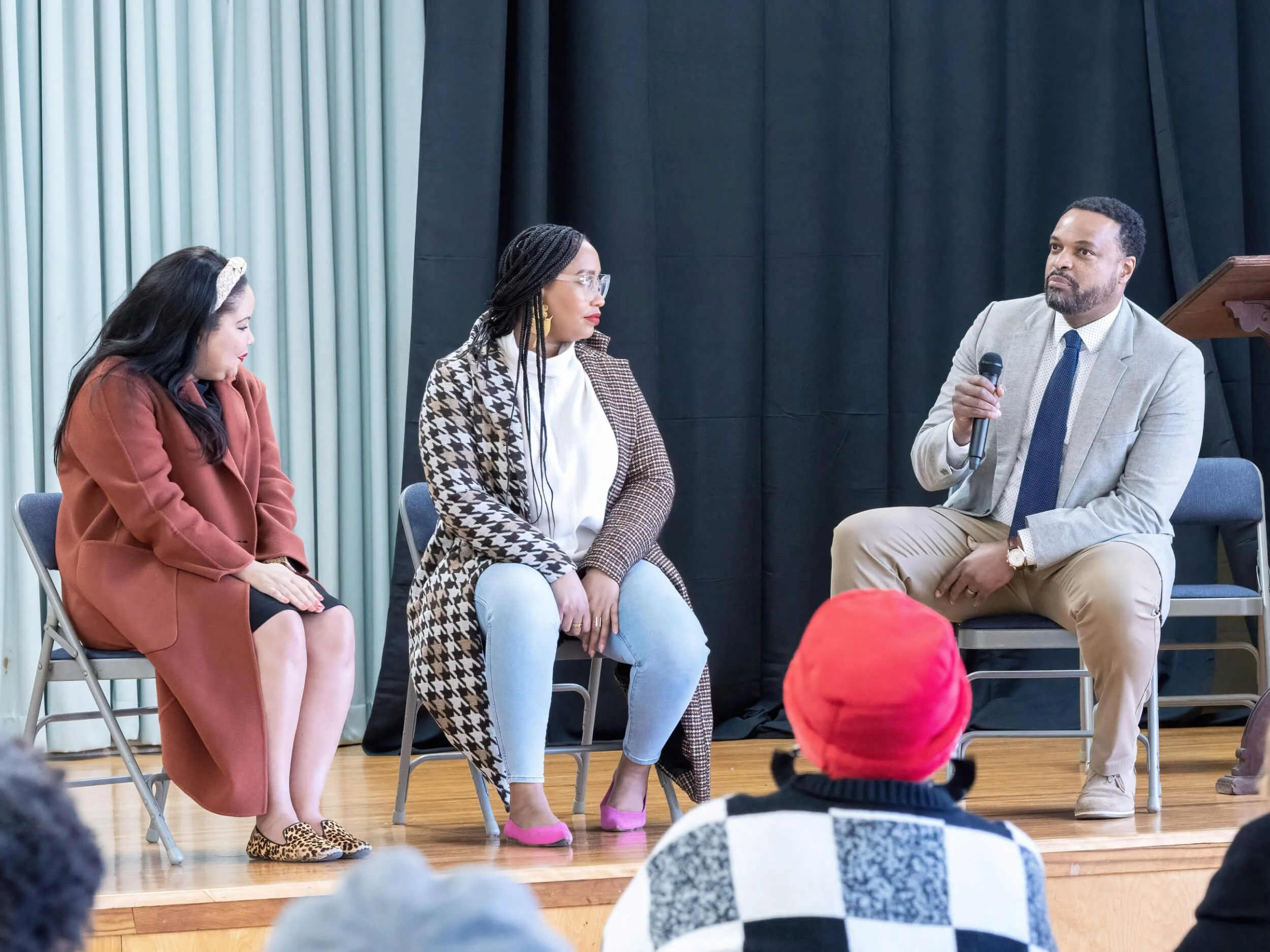 A panel discussion with three women and one man seated on stage, with an audience watching. The man is holding a microphone, and the panelists are engaged in conversation.