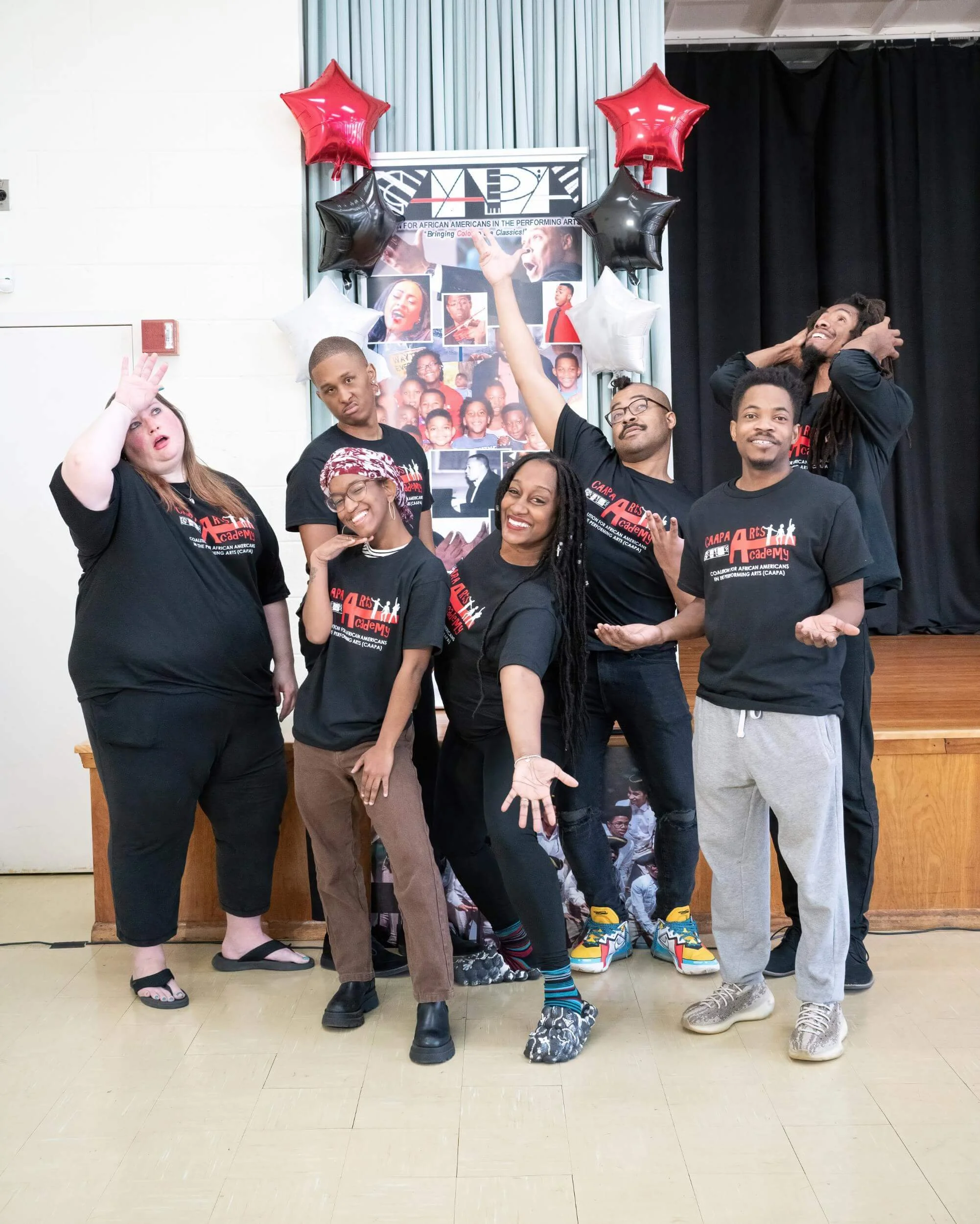 Group of seven people posing together in a room with a banner, balloons, and photos in the background. They are wearing matching black T-shirts with red and white text.