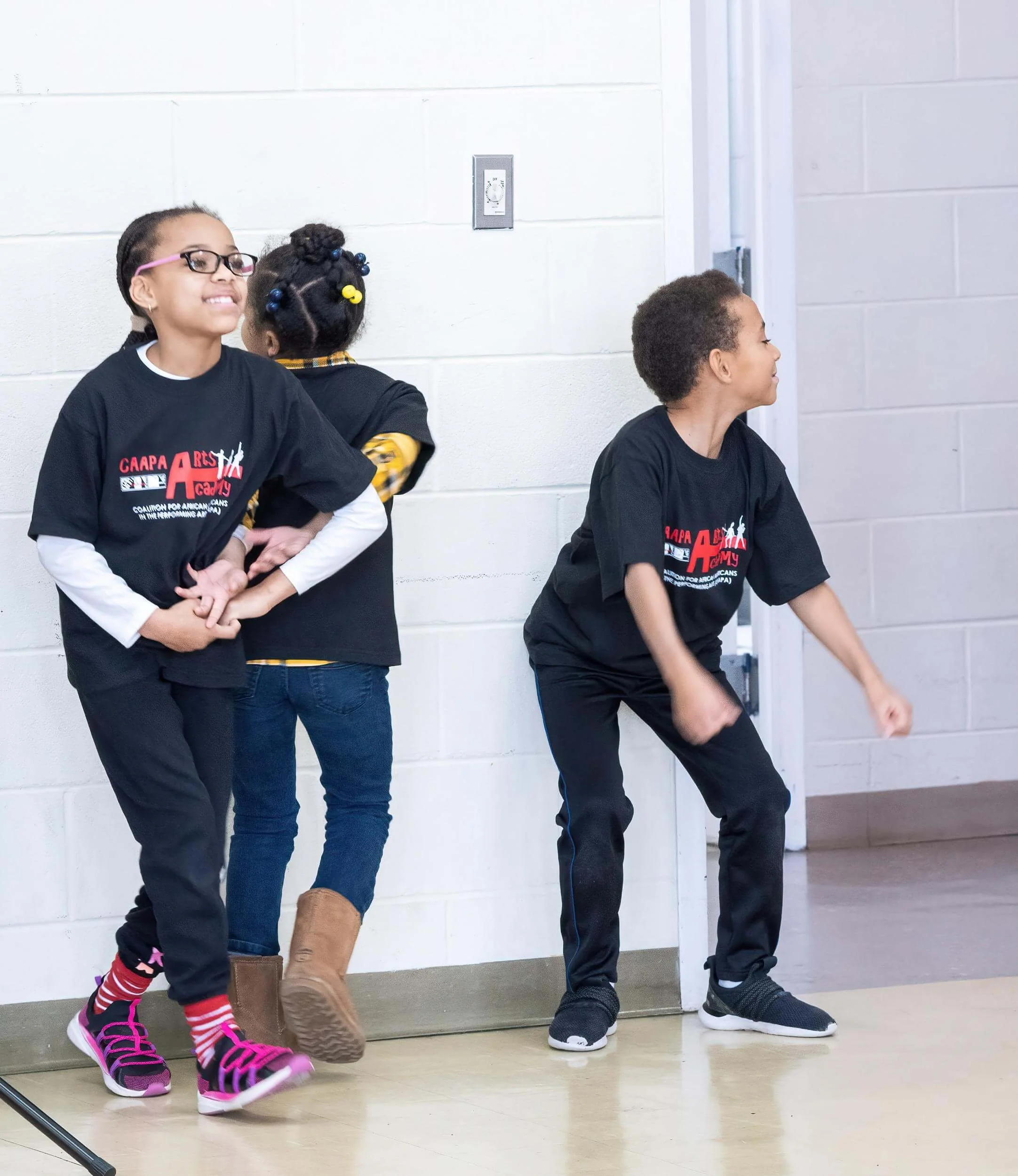 Three children standing against a white wall, wearing black t-shirts, one girl with glasses and two boys, one smiling and the other with fists clenched, appearing to dance or perform a move.