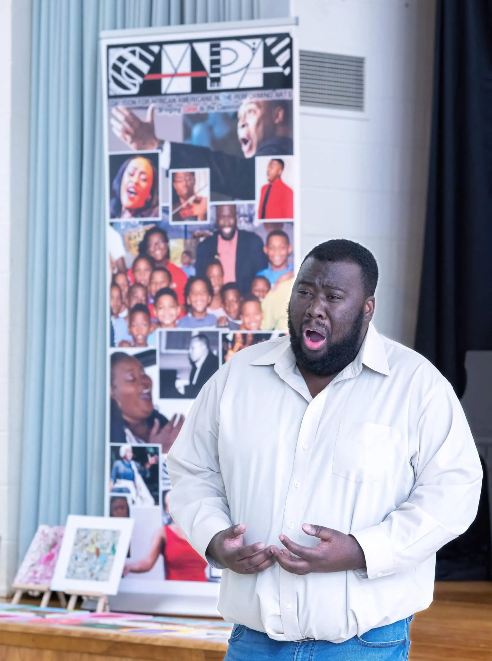 A man singing passionately in front of a collage of photographs, including a group photo of children and pictures of performers.