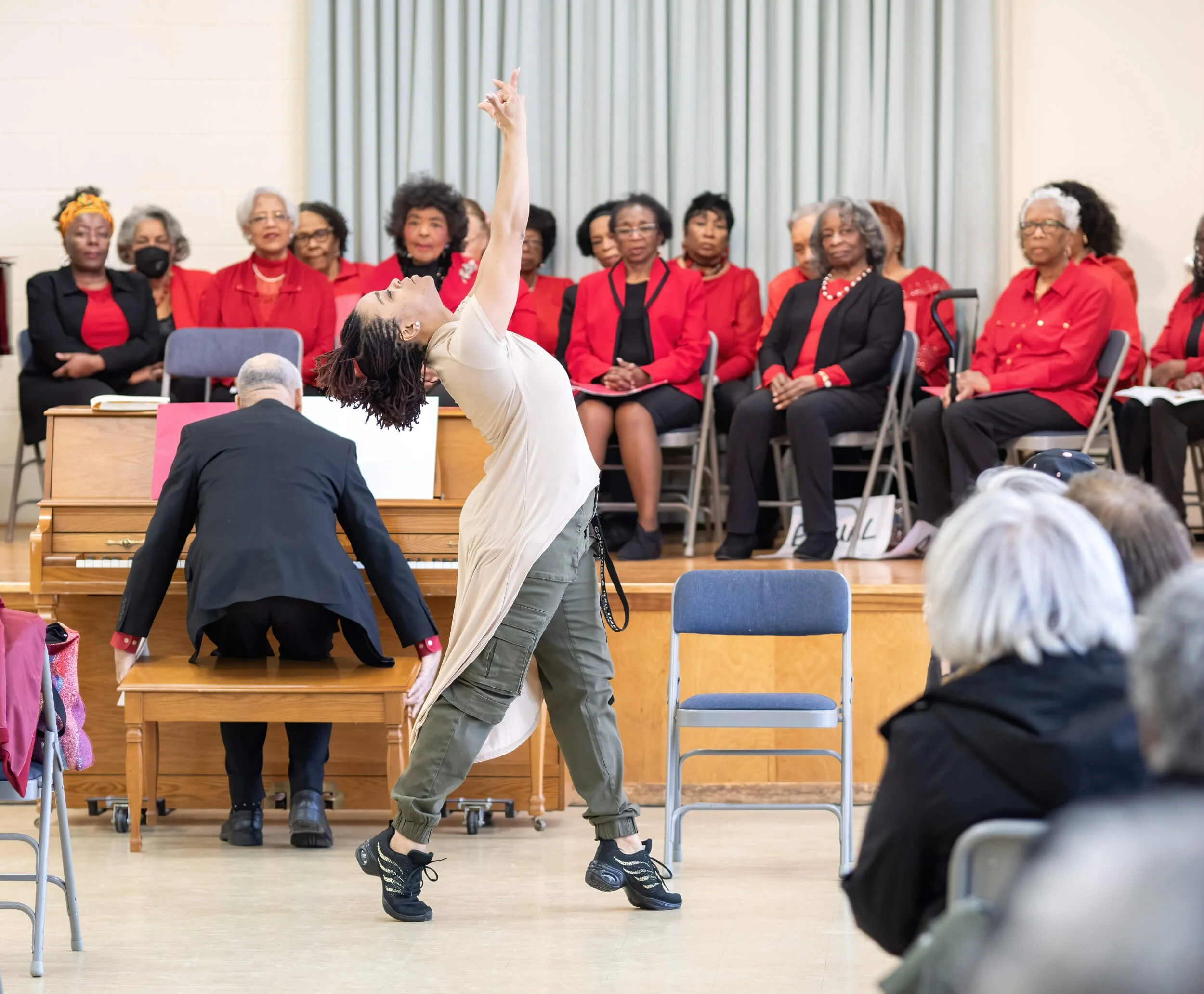 A woman dancing expressively on stage with a group of women seated behind her, some in red attire, observing the performance.