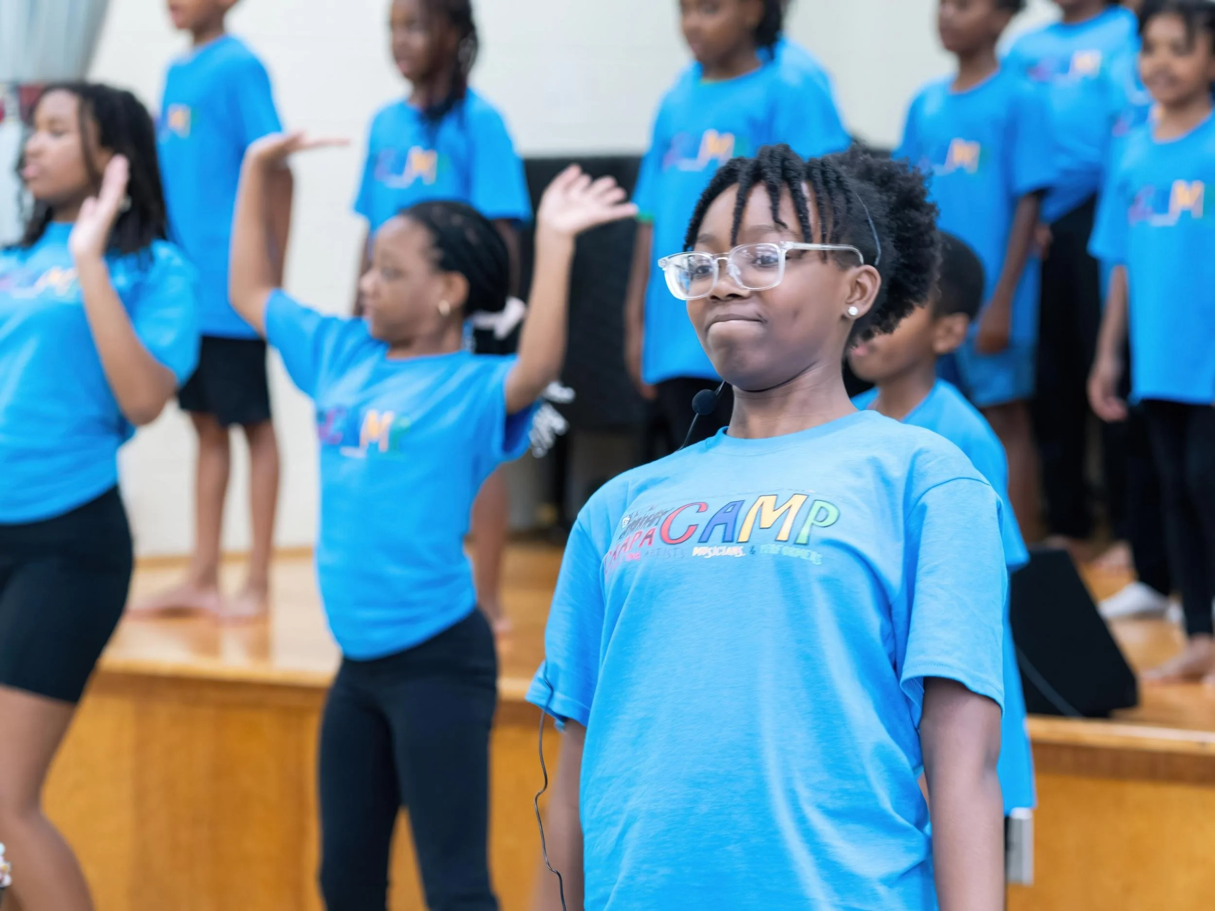 A group of children in blue t-shirts participating in a choir or singing performance, with some raising their hands and others standing on a stage.