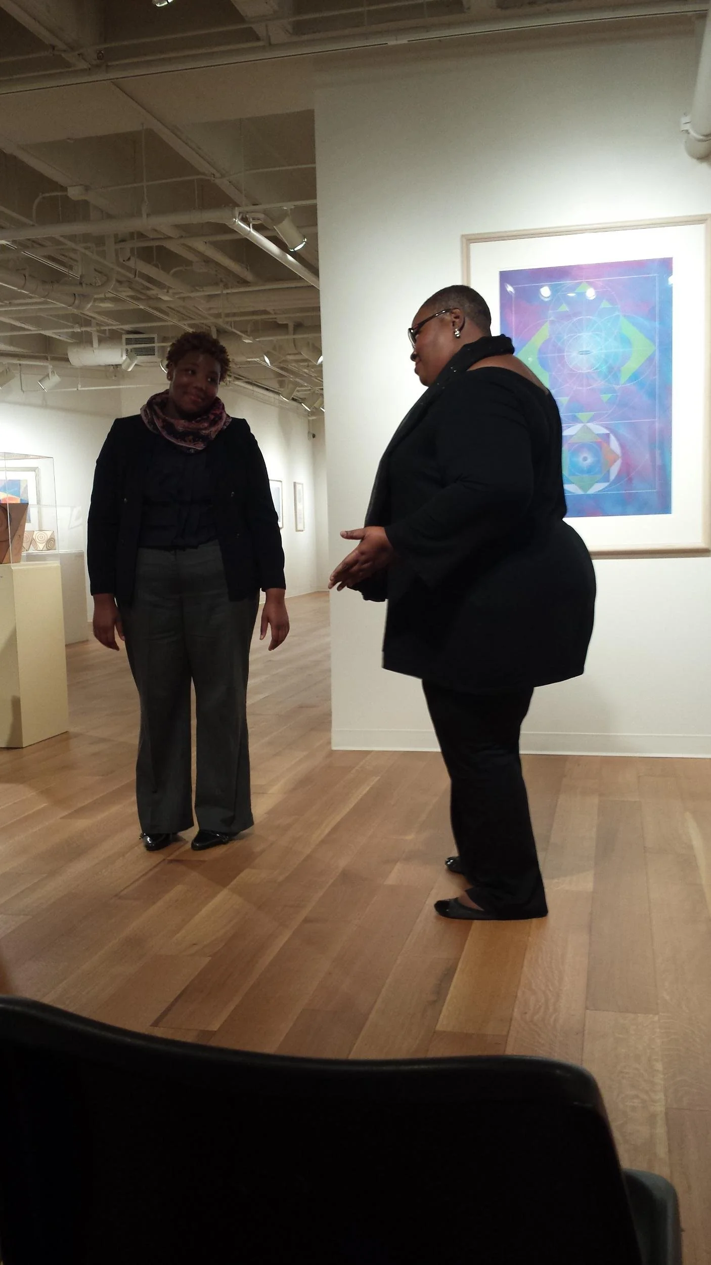 Two women having a conversation in an art gallery. One woman is wearing a black jacket and the other is dressed in all black.